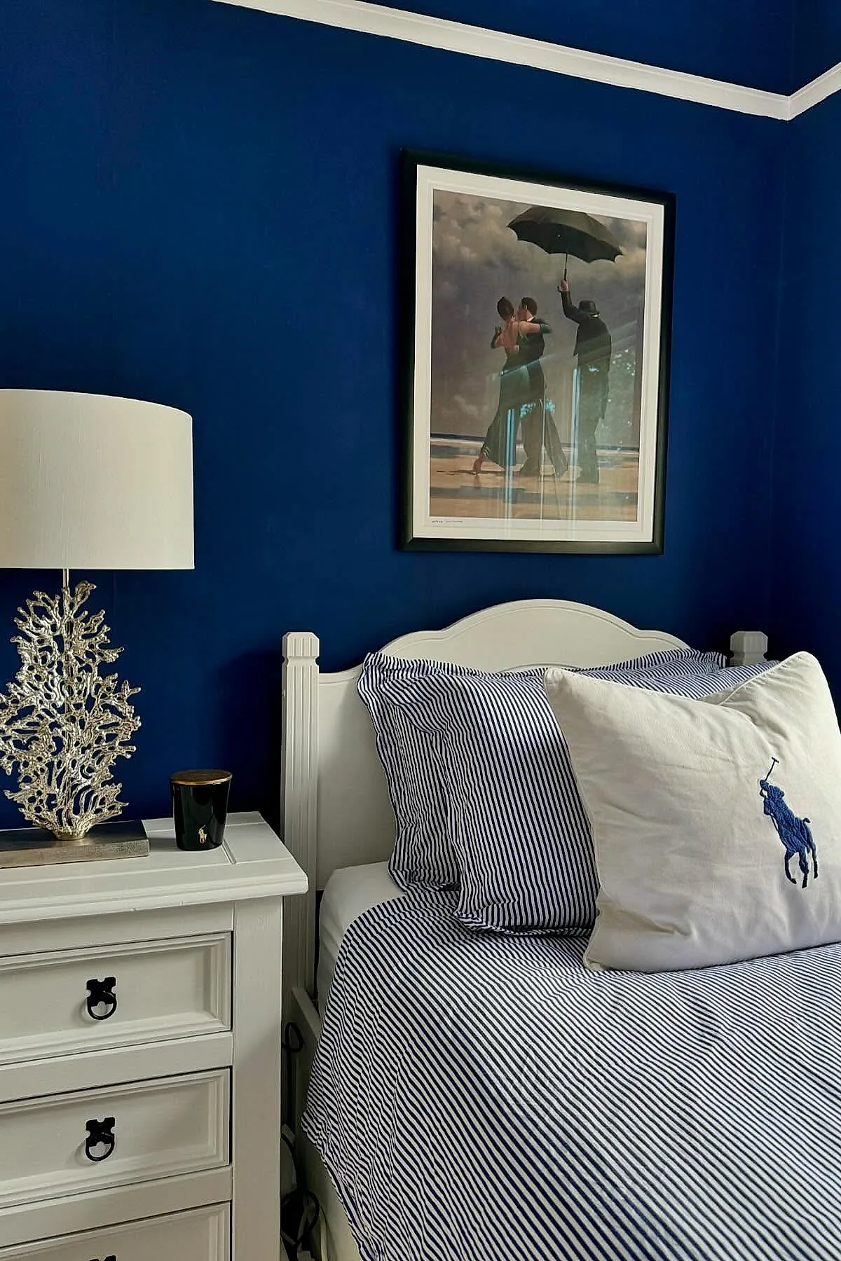 bedroom with deep navy blue walls, white wooden bed with navy and white striped bedding, large cream pillow embroidered with blue polo player, white nightstand holding silver coral-shaped lamp and small black candle, framed artwork above bed showing people on beach under umbrella