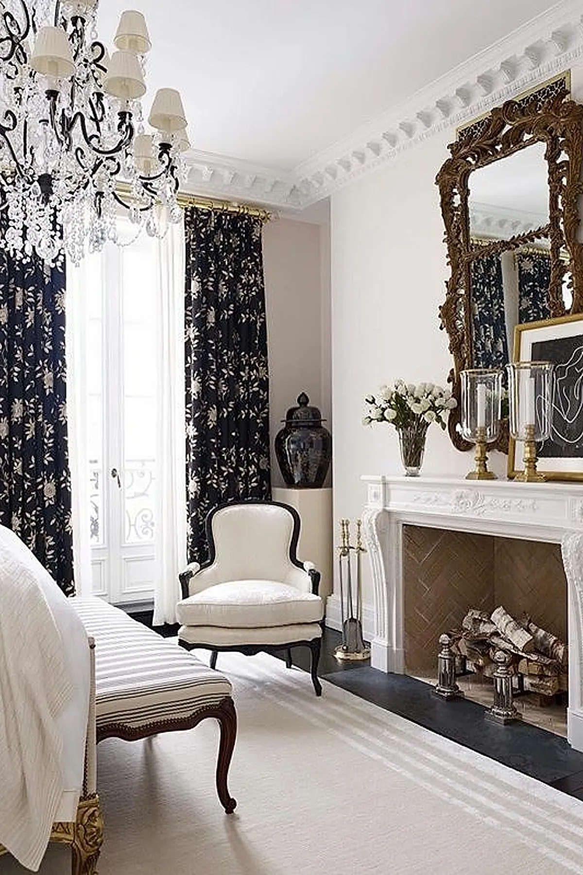 bright bedroom featuring large ornate gold framed mirror above white fireplace, black floral curtains on tall windows, crystal chandelier, carved wooden bench with striped cushion, upholstered armchair with dark frame