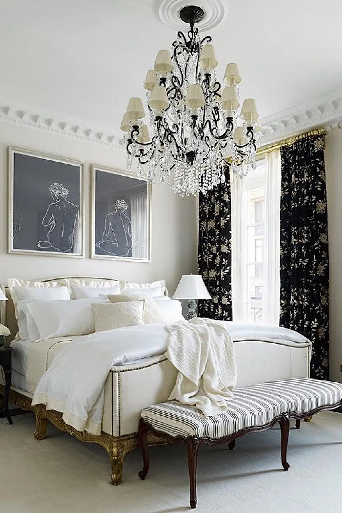 traditional bedroom with white walls, black floral curtains, crystal chandelier, upholstered white bed with gold trim, striped cushioned bench at foot of bed, two framed minimalist line drawings above headboard