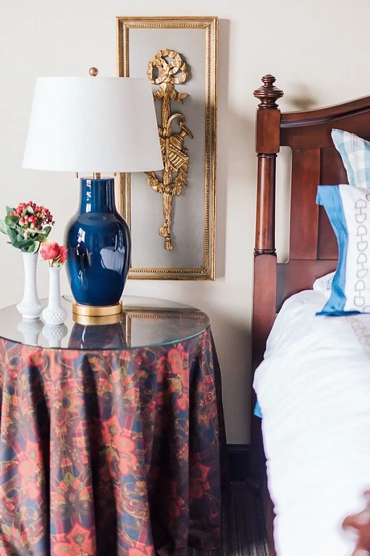 side table covered in dark red and blue patterned fabric with glass top holding a blue ceramic lamp with white shade, gold decorative wall art behind, part of wooden bed frame visible on right