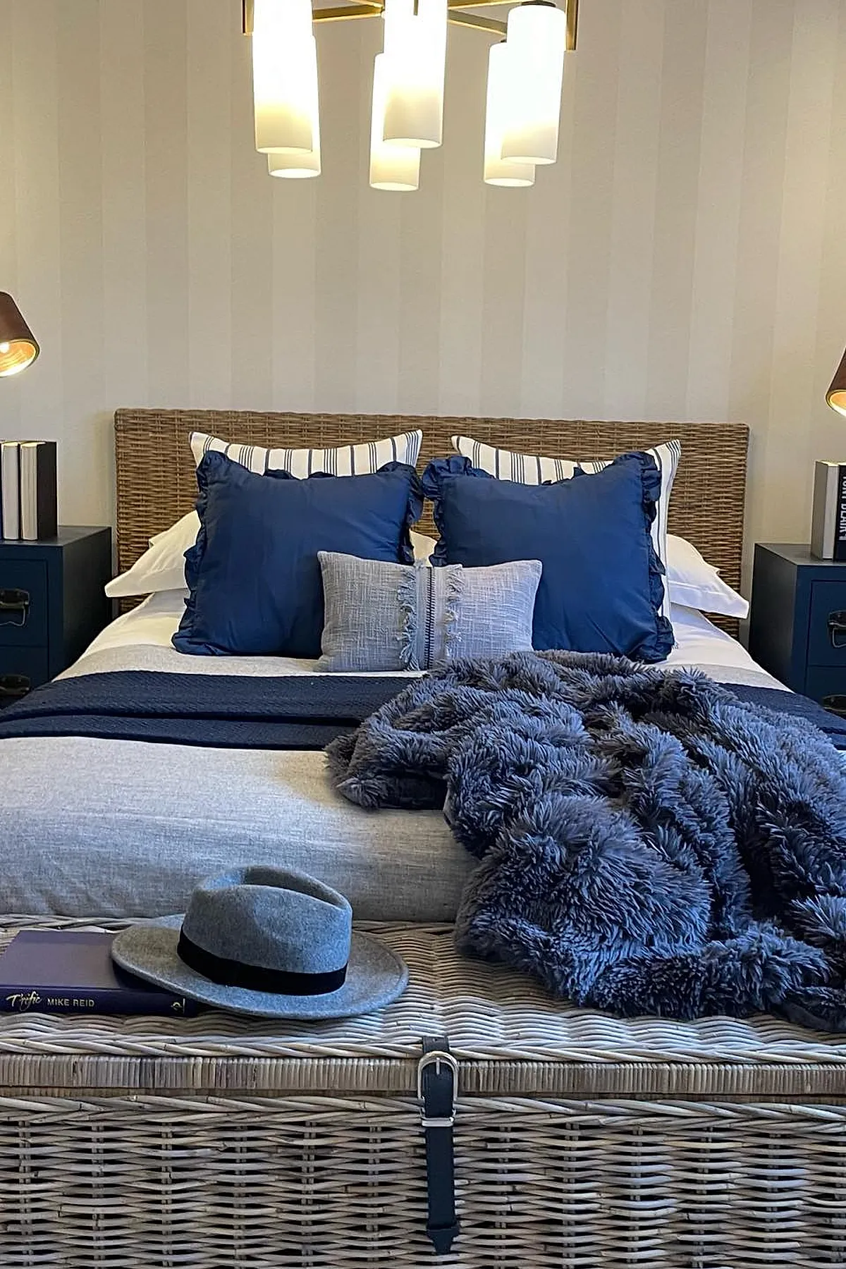 bed with woven rattan headboard, white bedding layered with blue striped and solid navy pillows, navy throw blanket, fluffy dark blue faux fur blanket, gray felt hat resting on woven storage bench at foot of bed
