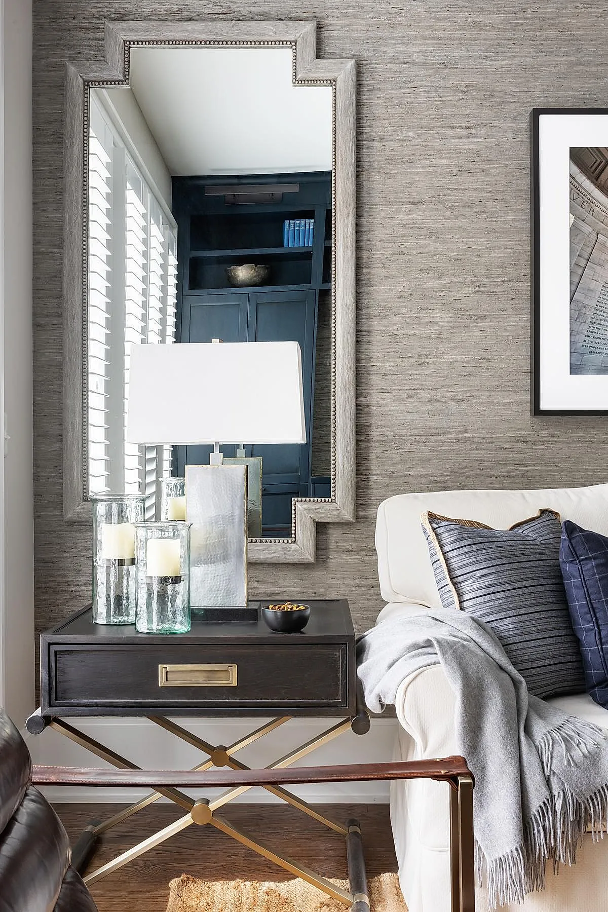 neutral textured wall with large stepped silver-framed mirror reflecting window shutters, black wooden side table with brass hardware holding two glass candle holders and a hammered metal lamp with white shade, white sofa with blue striped pillows and gray throw blanket
