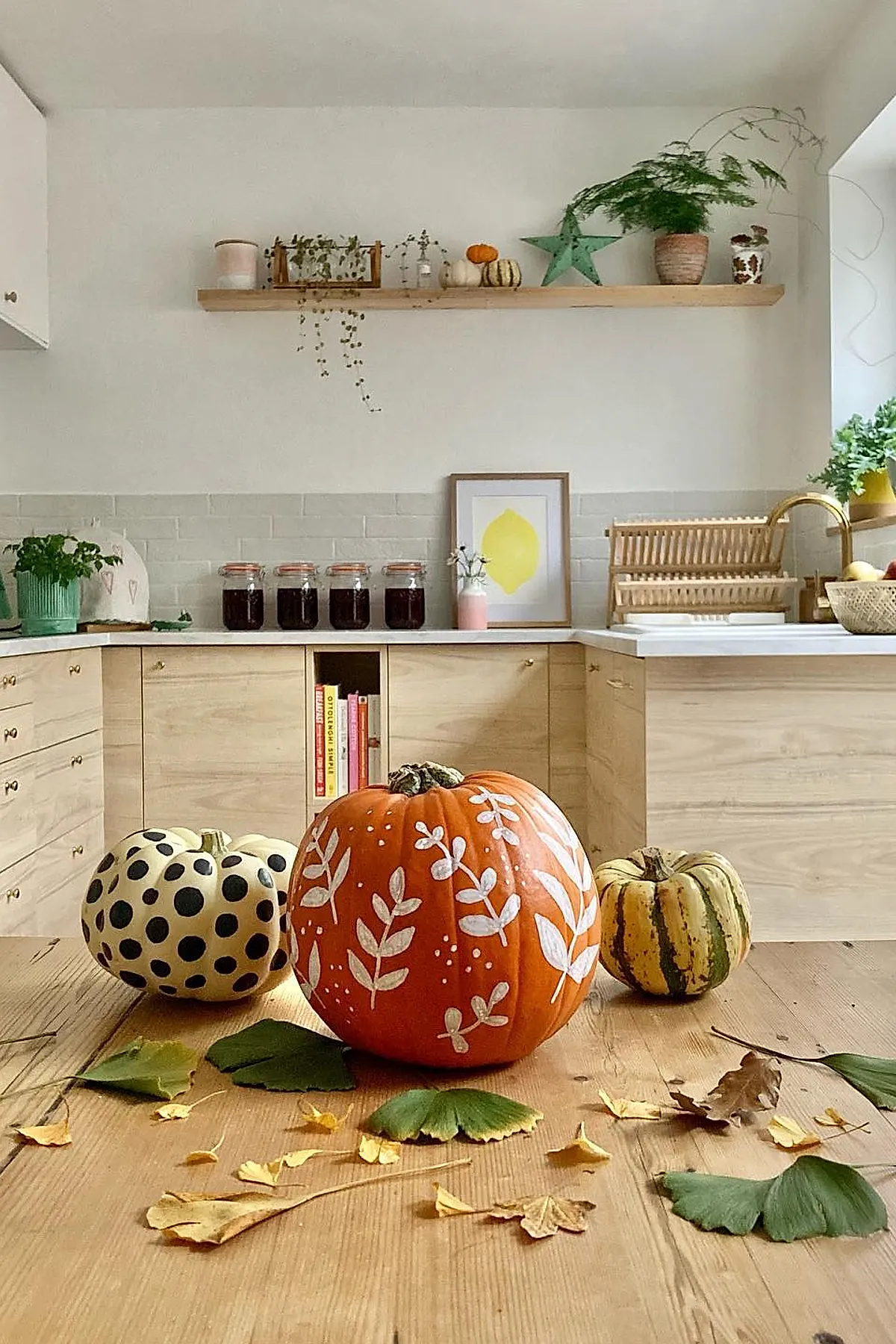 three pumpkins on wooden table in light kitchen one orange pumpkin painted with white leaves one cream pumpkin with black polka dots one small striped yellow green pumpkin scattered dry leaves