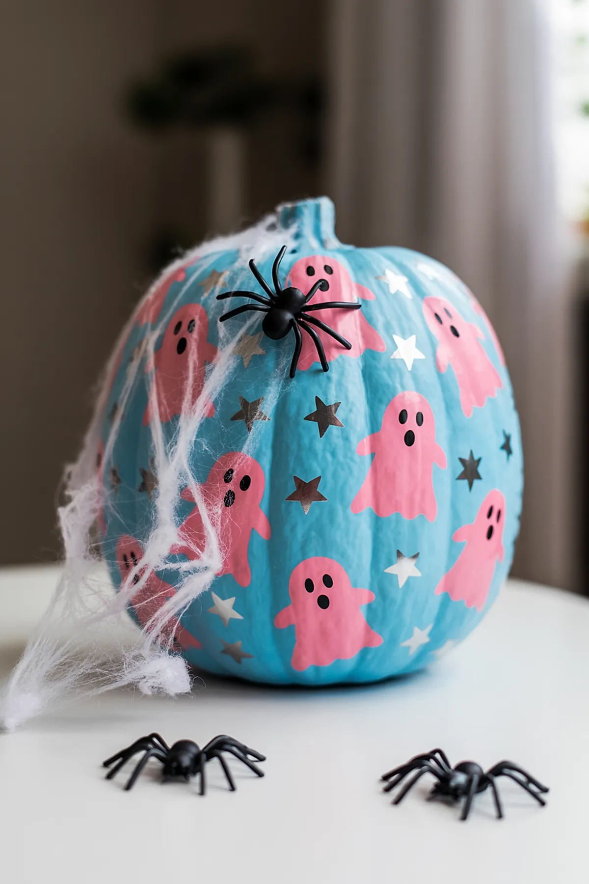 blue painted pumpkin decorated with pink ghost shapes and silver star stickers, adorned with white fake spider web and black plastic spiders on white surface
