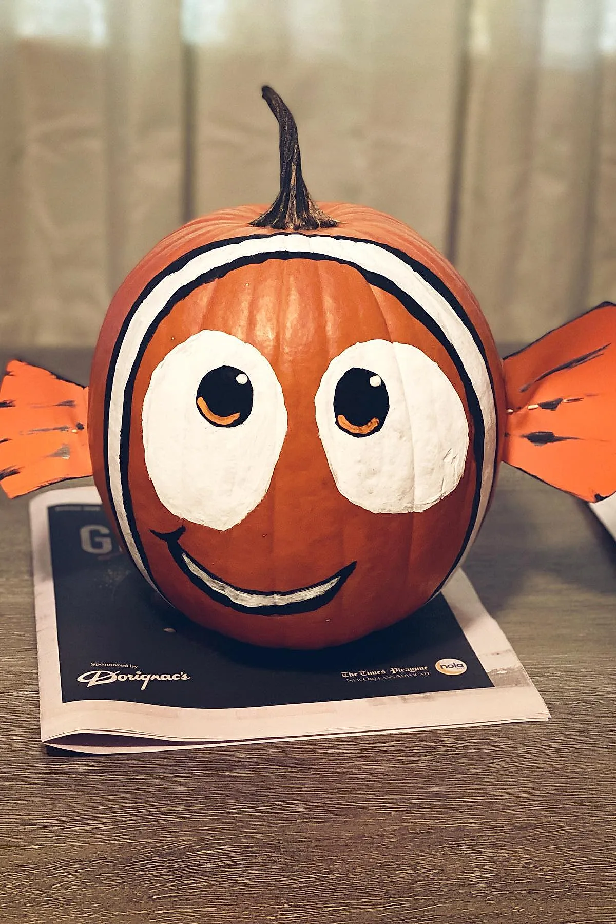 orange pumpkin painted with large white eyes outlined in black and black pupils with smiling mouth painted on front orange paper fins attached on each side