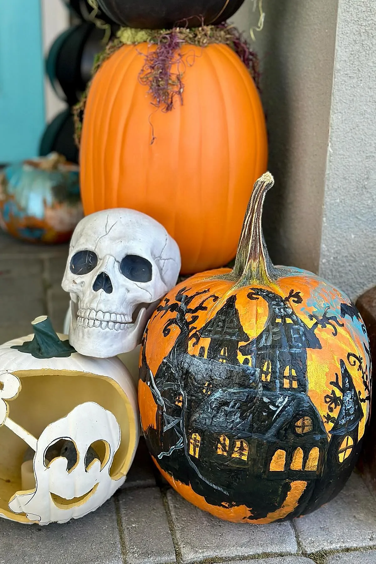 orange pumpkin hand painted with black haunted house scene next to white ceramic skull and white pumpkin carved with smiling ghost face on outdoor stone step