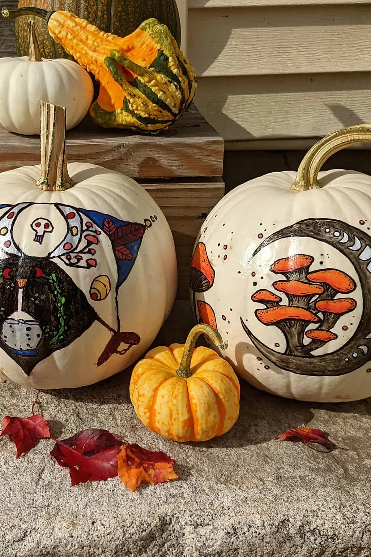 two white pumpkins decorated with colorful mushroom illustrations and small skull and crescent moon motifs placed on stone step with autumn leaves