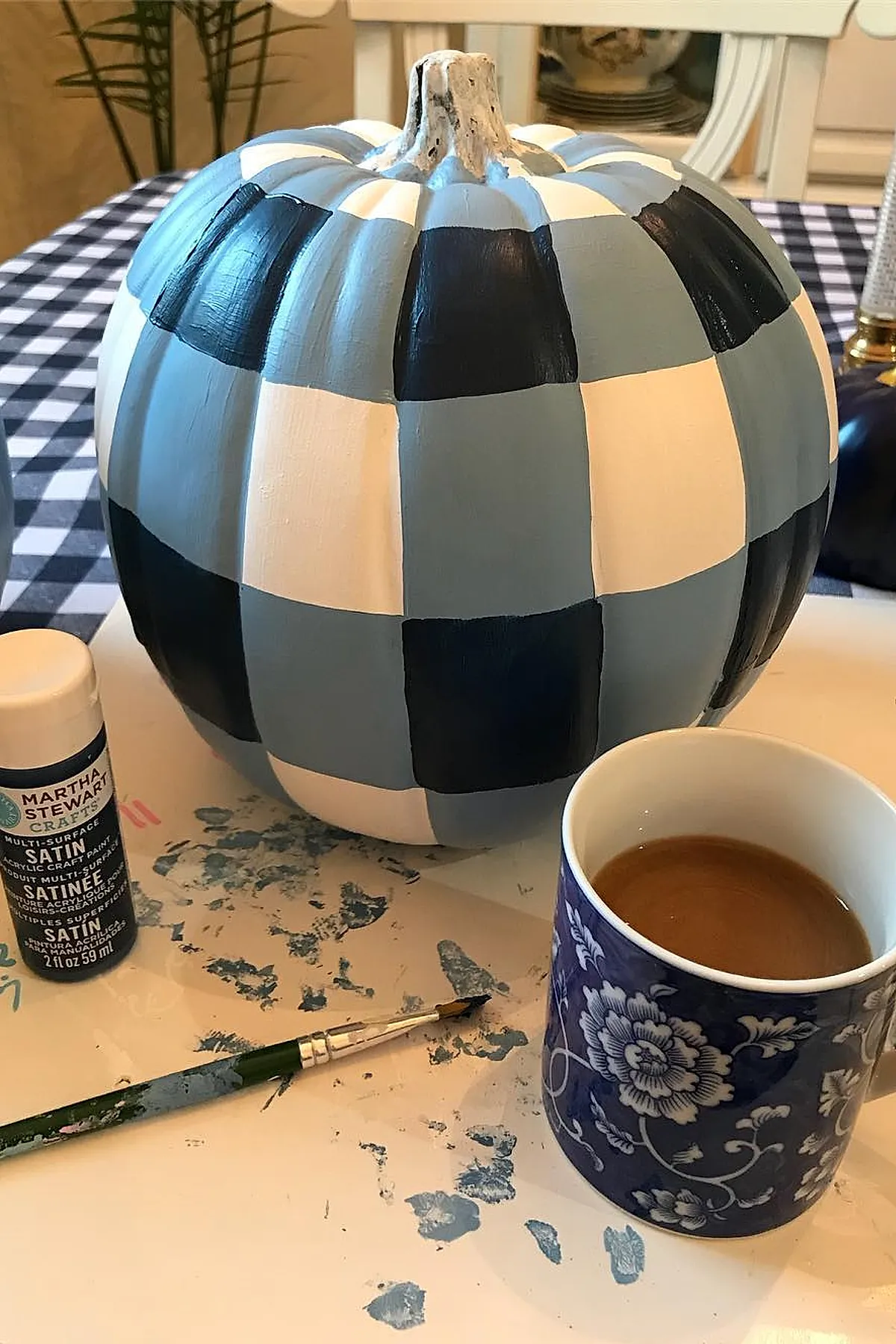 pumpkin painted in alternating blue, white, and black squares in buffalo check pattern sitting on table next to paintbrush, paint bottle, and floral blue coffee cup