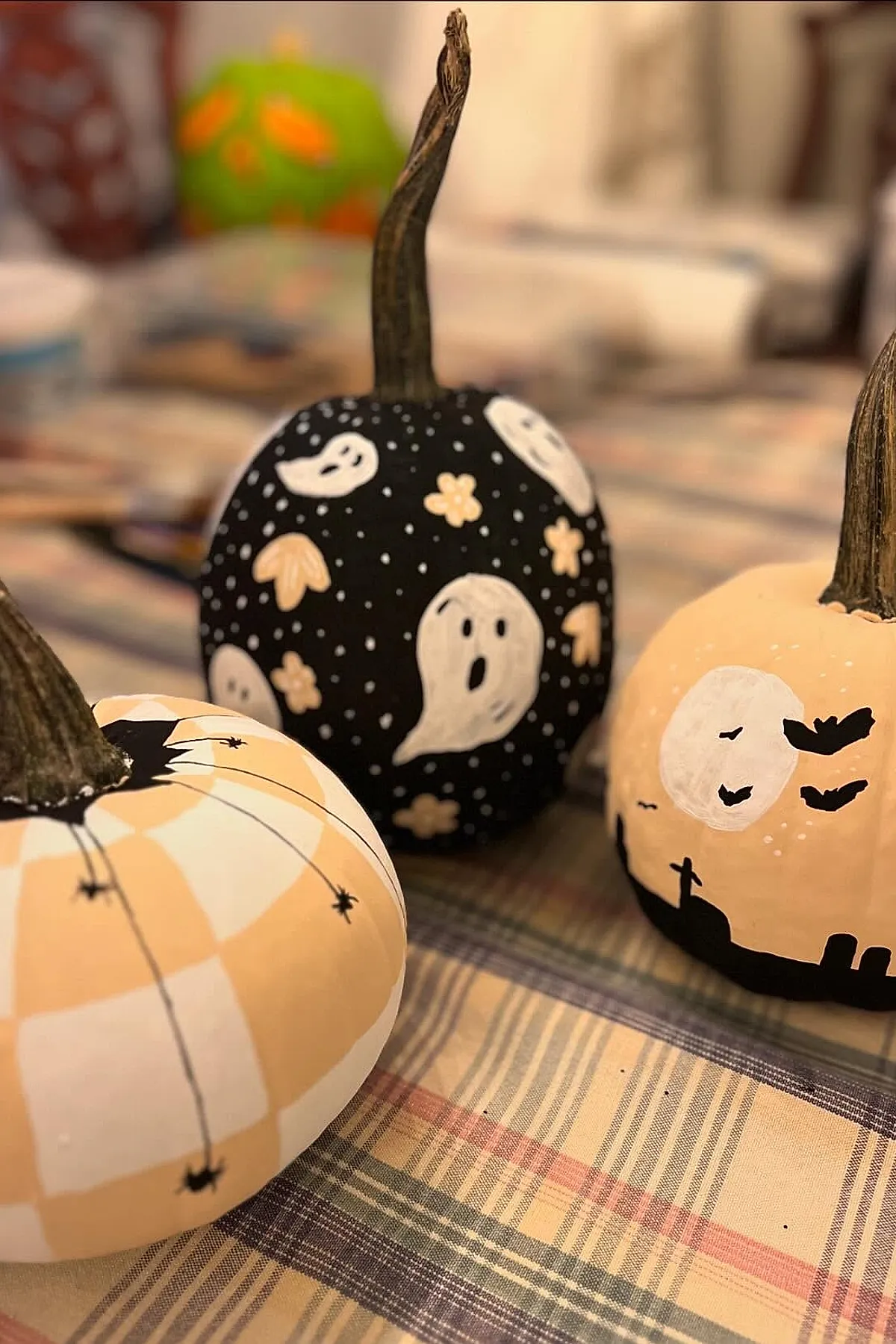 three small pumpkins painted with Halloween designs including white ghosts, black bats, spider webs, and spiders on a plaid fabric surface
