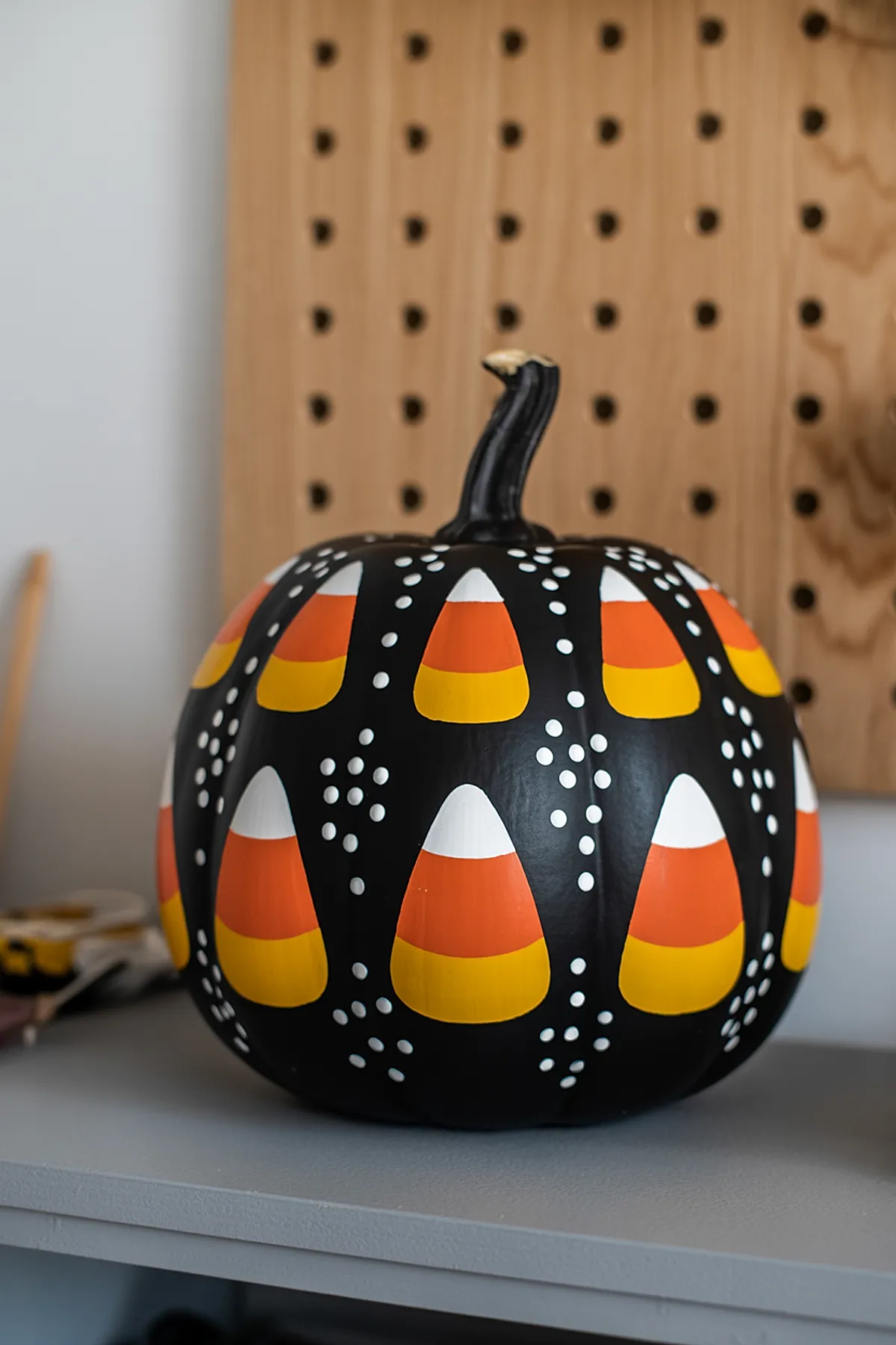 black painted pumpkin decorated with repeated candy corn shapes in white, orange, and yellow surrounded by small white dots sitting on gray surface with wooden pegboard background