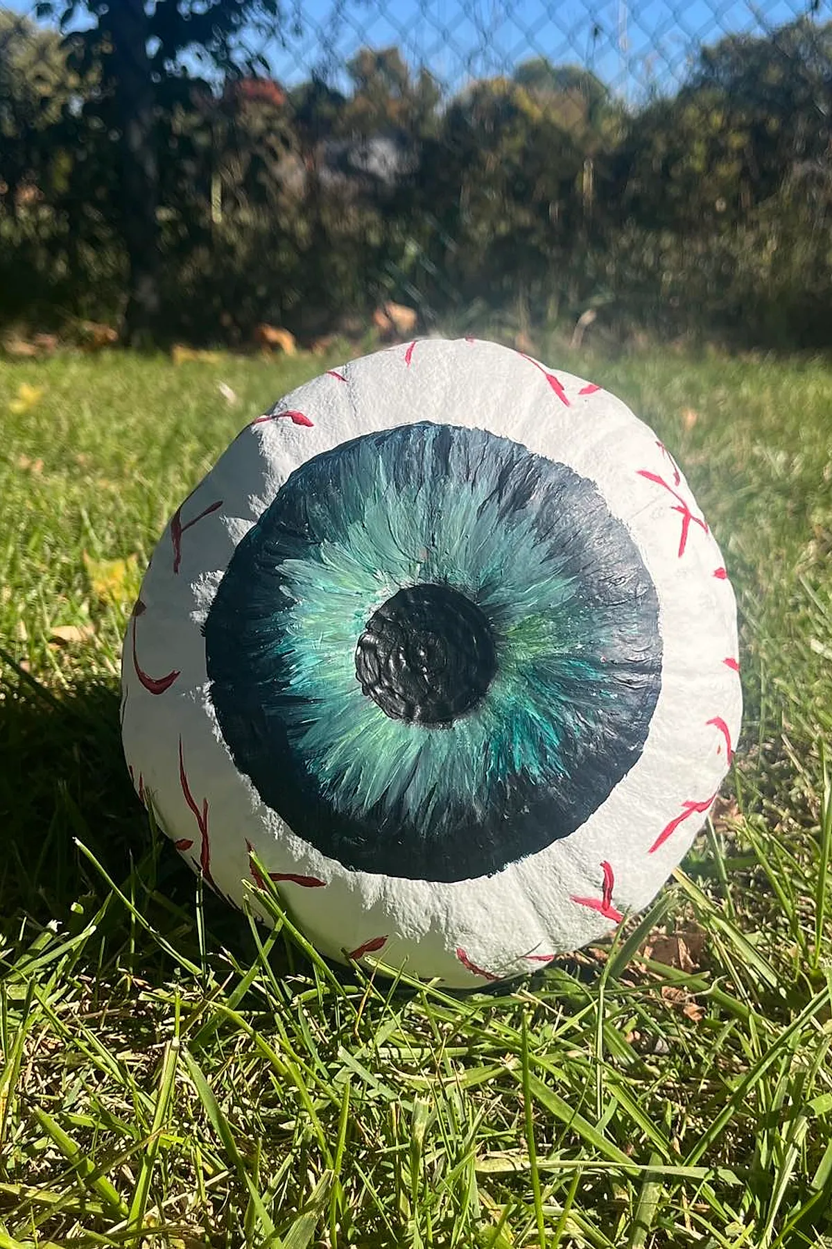 white pumpkin painted with large realistic blue and green iris and black pupil with red veins painted on surface placed on green grass