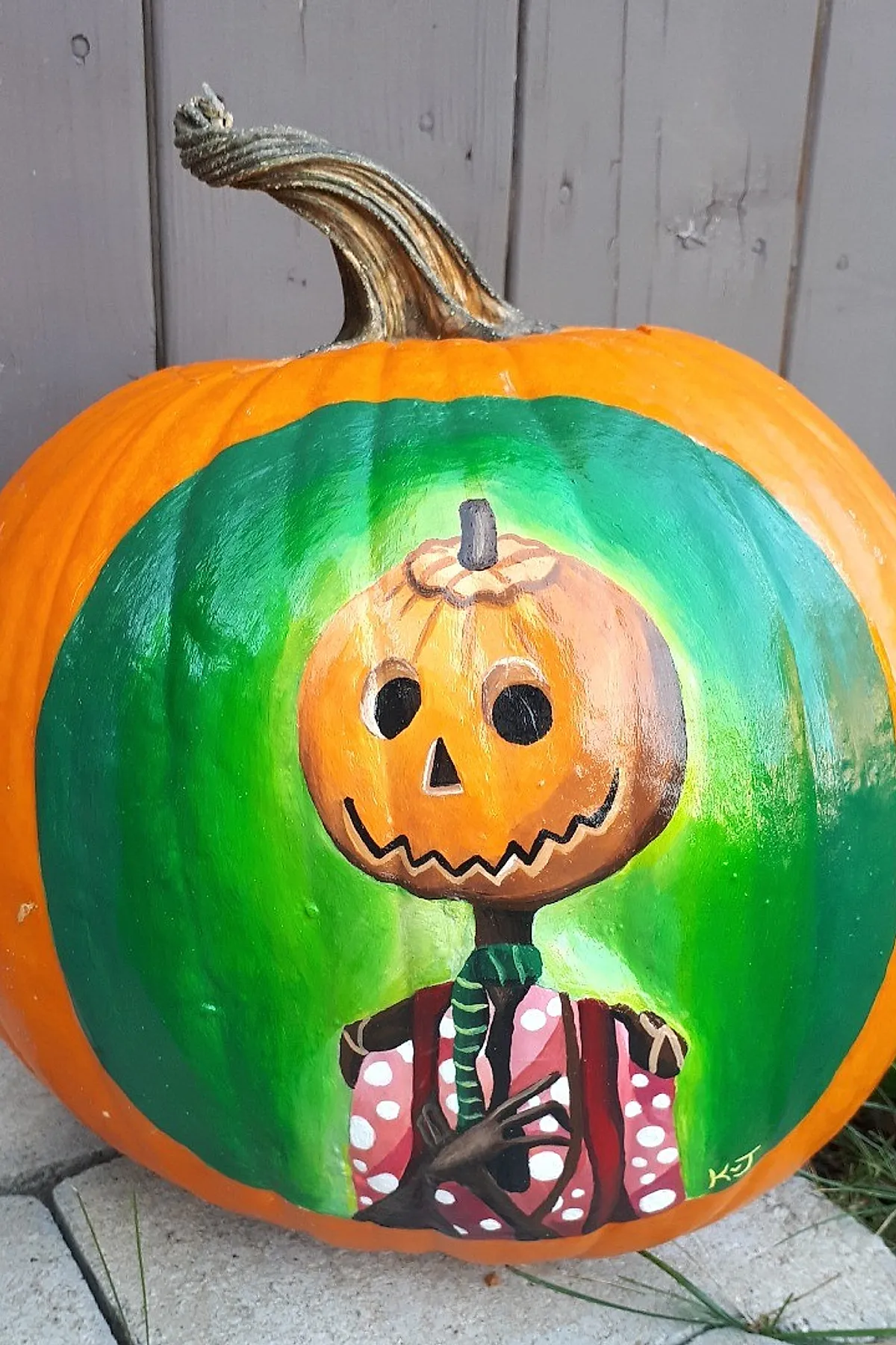 orange pumpkin painted with smiling jack o lantern face wearing pink polka dot shirt and green striped scarf on green background