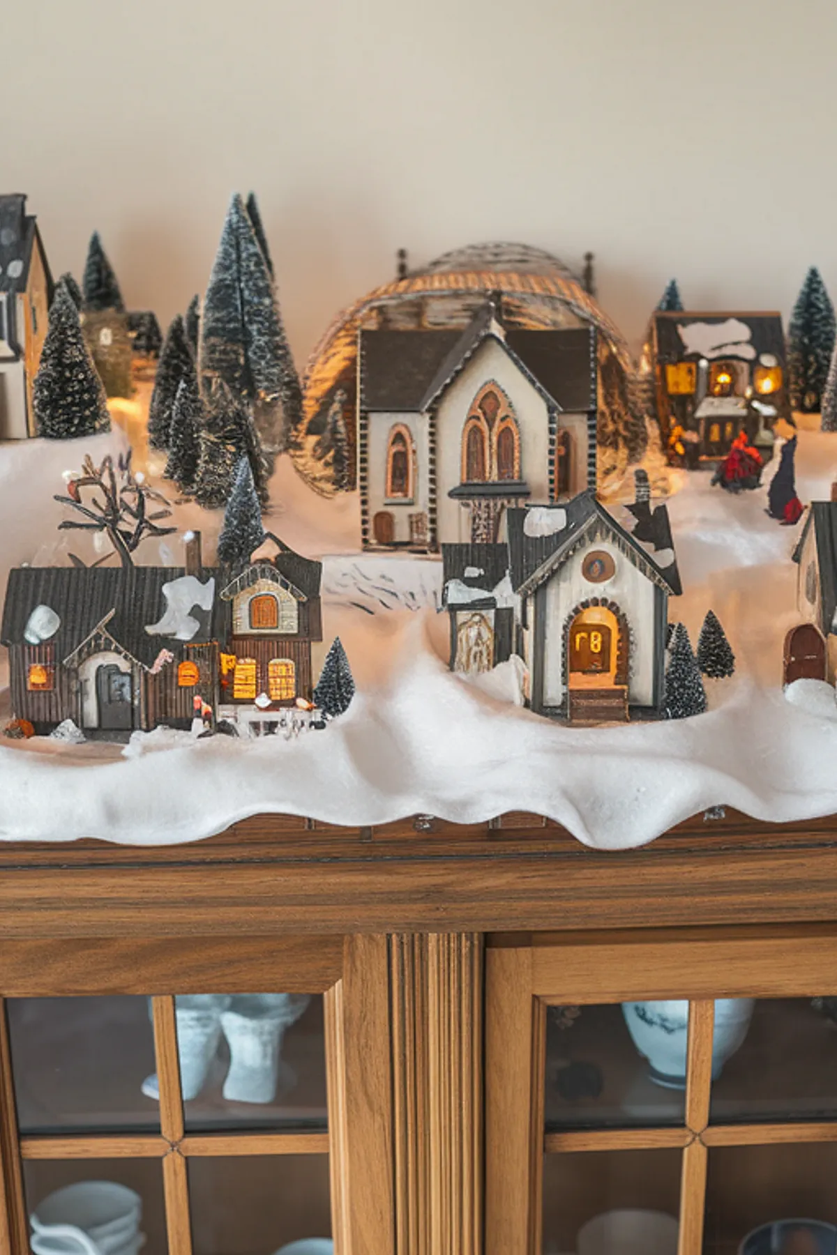 Meticulously crafted miniature village with illuminated houses and snow-covered trees, displayed on a wooden cabinet against a white backdrop.