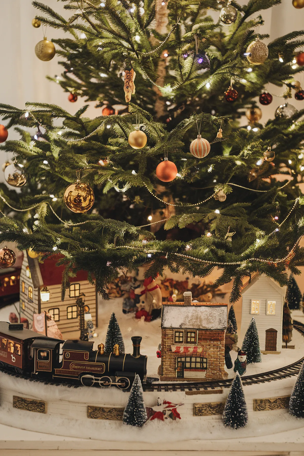 Decorated Christmas tree with gold, red, and silver ornaments, surrounded by a miniature village and dark red train on snowy ground.