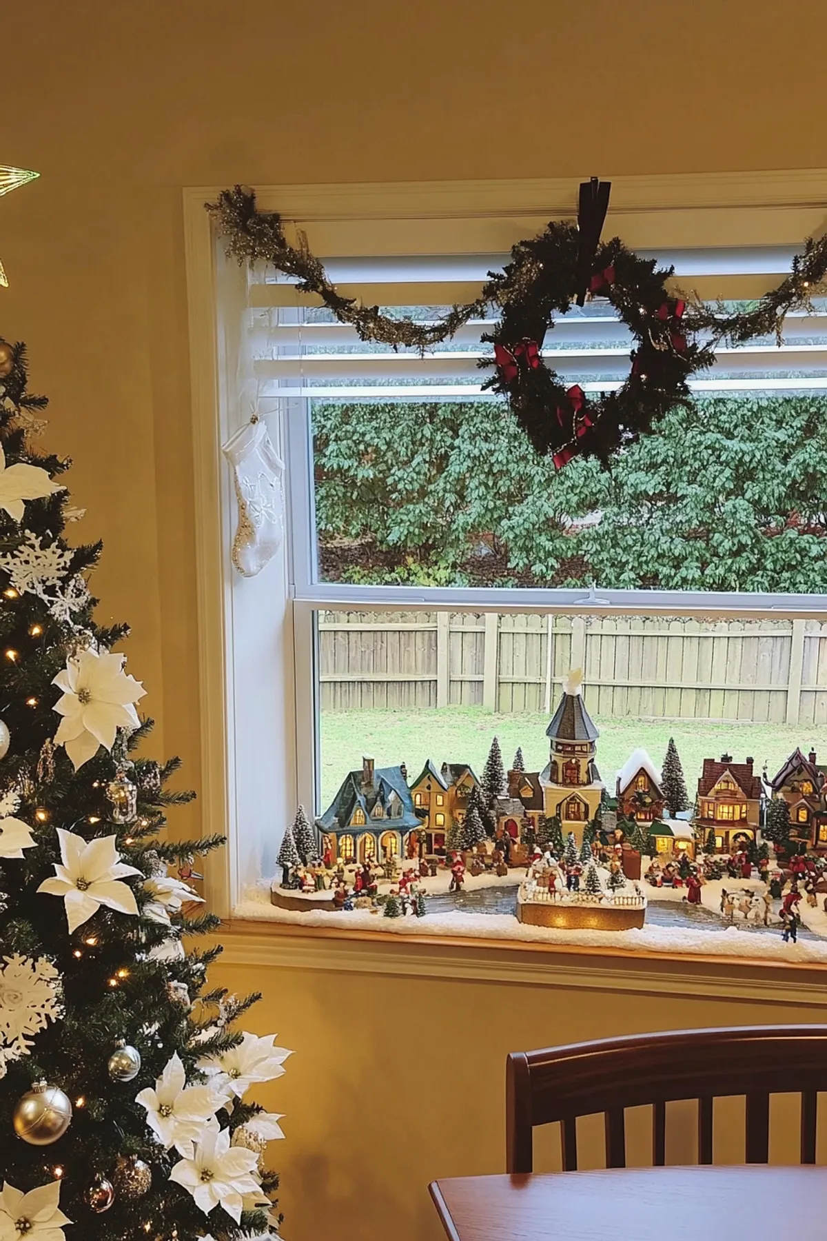 Christmas tree with silver ornaments and poinsettias beside a miniature village scene, warm yellow walls, and a festive wreath.