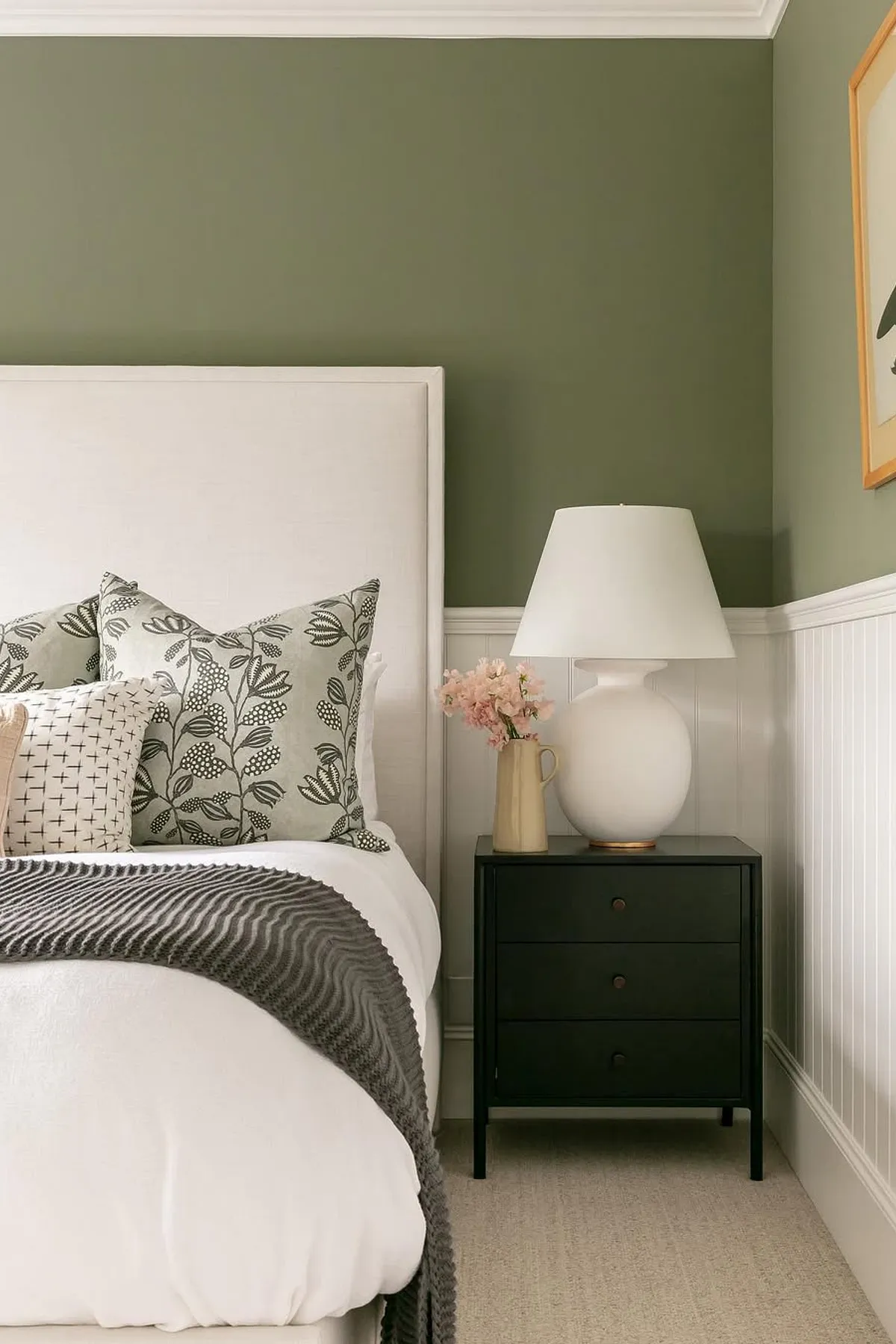 A stylish bedroom with a white bed, green walls, gray and white pillows, and a black nightstand with pink flowers.