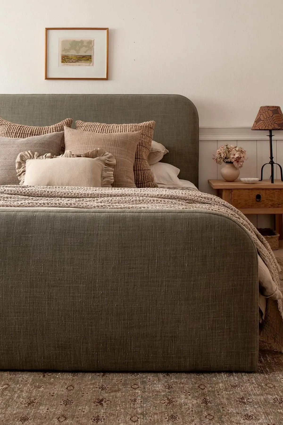 A serene bedroom with a muted green bed, neutral pillows, patterned lamp, framed art, and a textured rug.