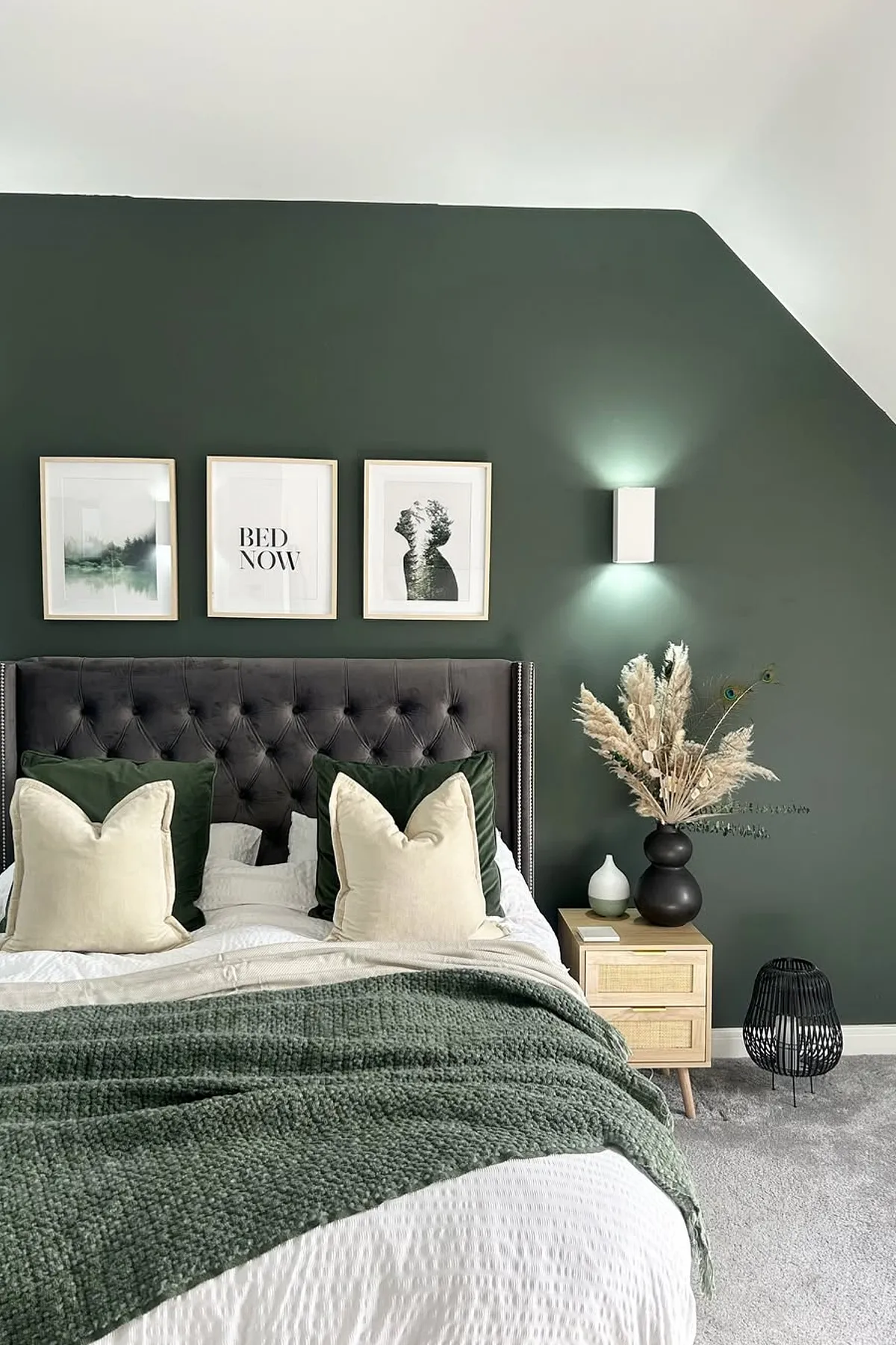 A cozy bedroom with a green accent wall, white bedding, framed art, wooden nightstand with pampas grass, and a modern lamp.