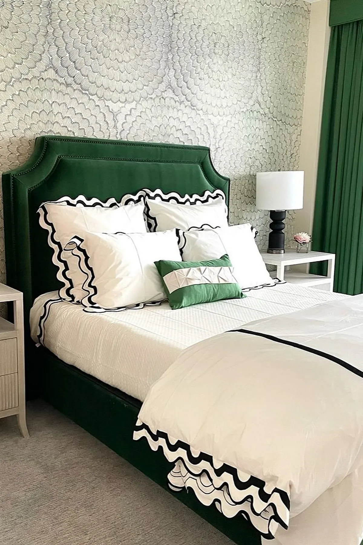 A bedroom featuring a green upholstered bed, floral wallpaper, white bedding, black lamps, and natural light from a large window.