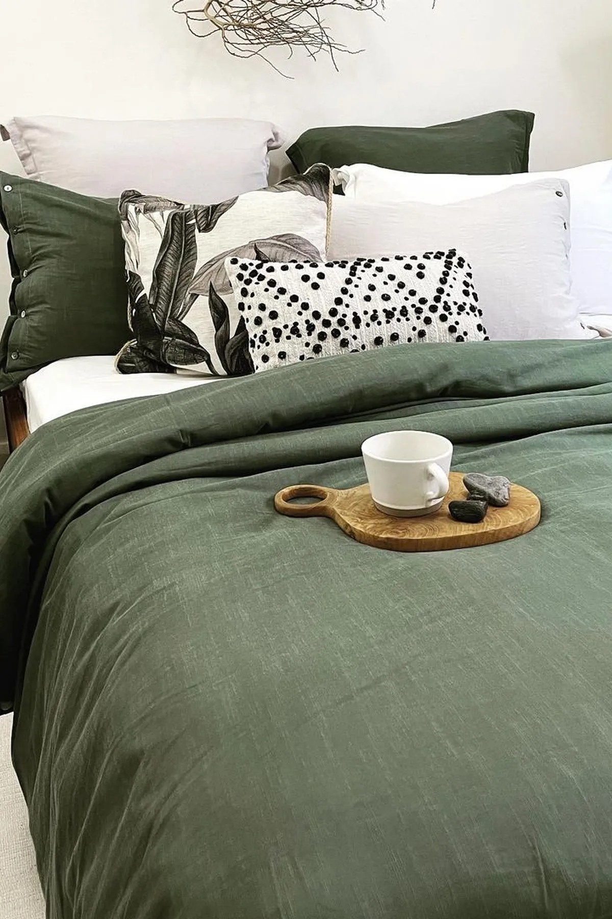 A cozy bedroom featuring a deep green duvet, decorative pillows, a wooden tray, and branch wall art above the bed.