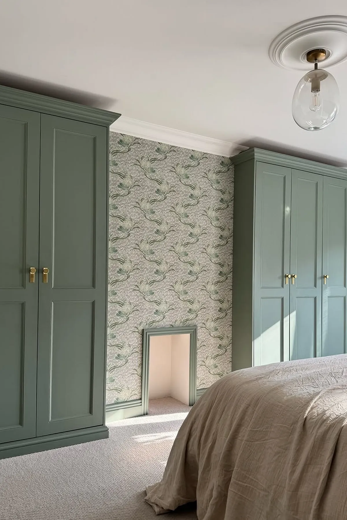 A bedroom with green wardrobes, floral wallpaper, a pendant light, and a window with white curtains allowing natural light.
