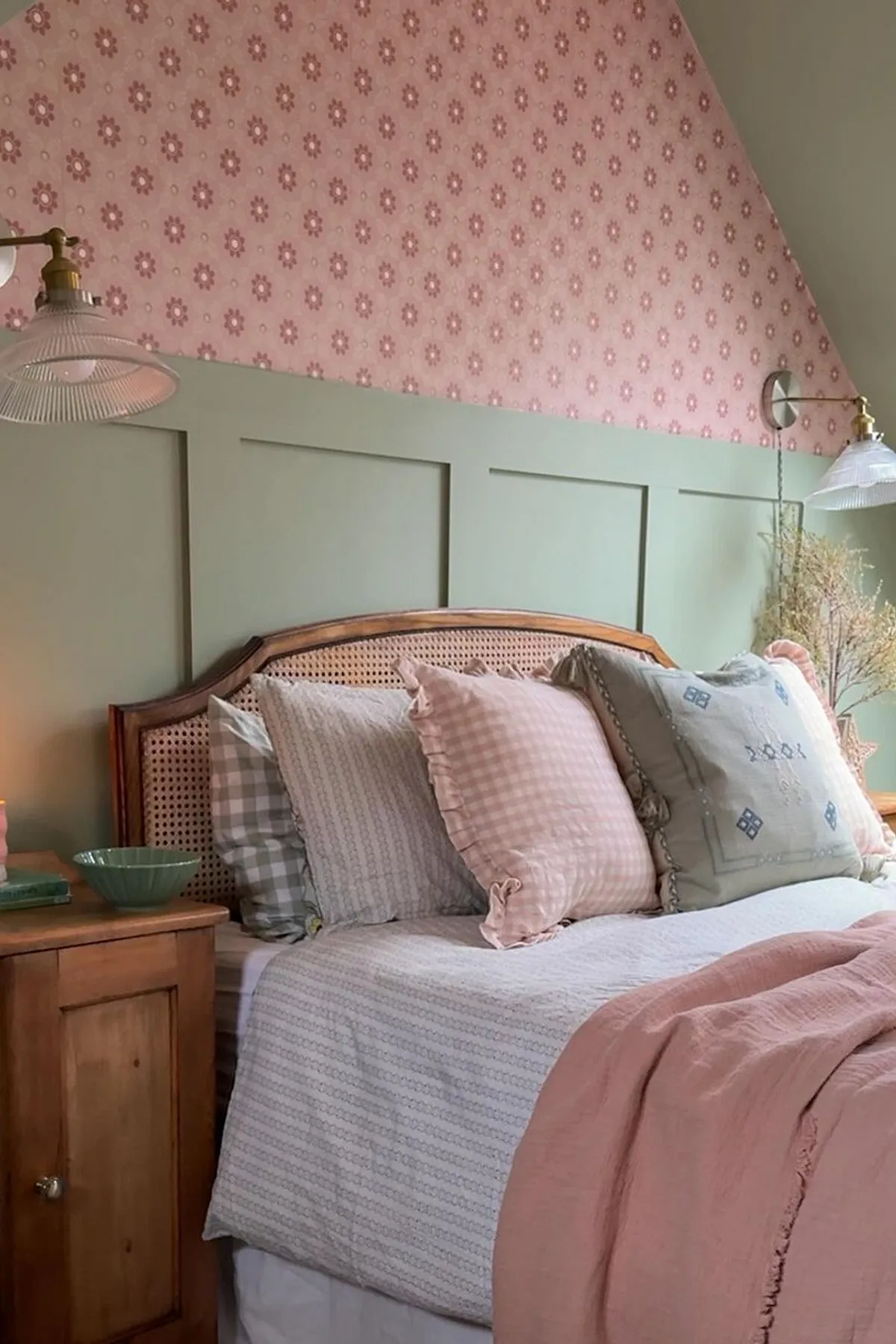 A cozy bedroom with a wooden bed, pink floral wall, hanging lamps, and a nightstand featuring a candle and book.