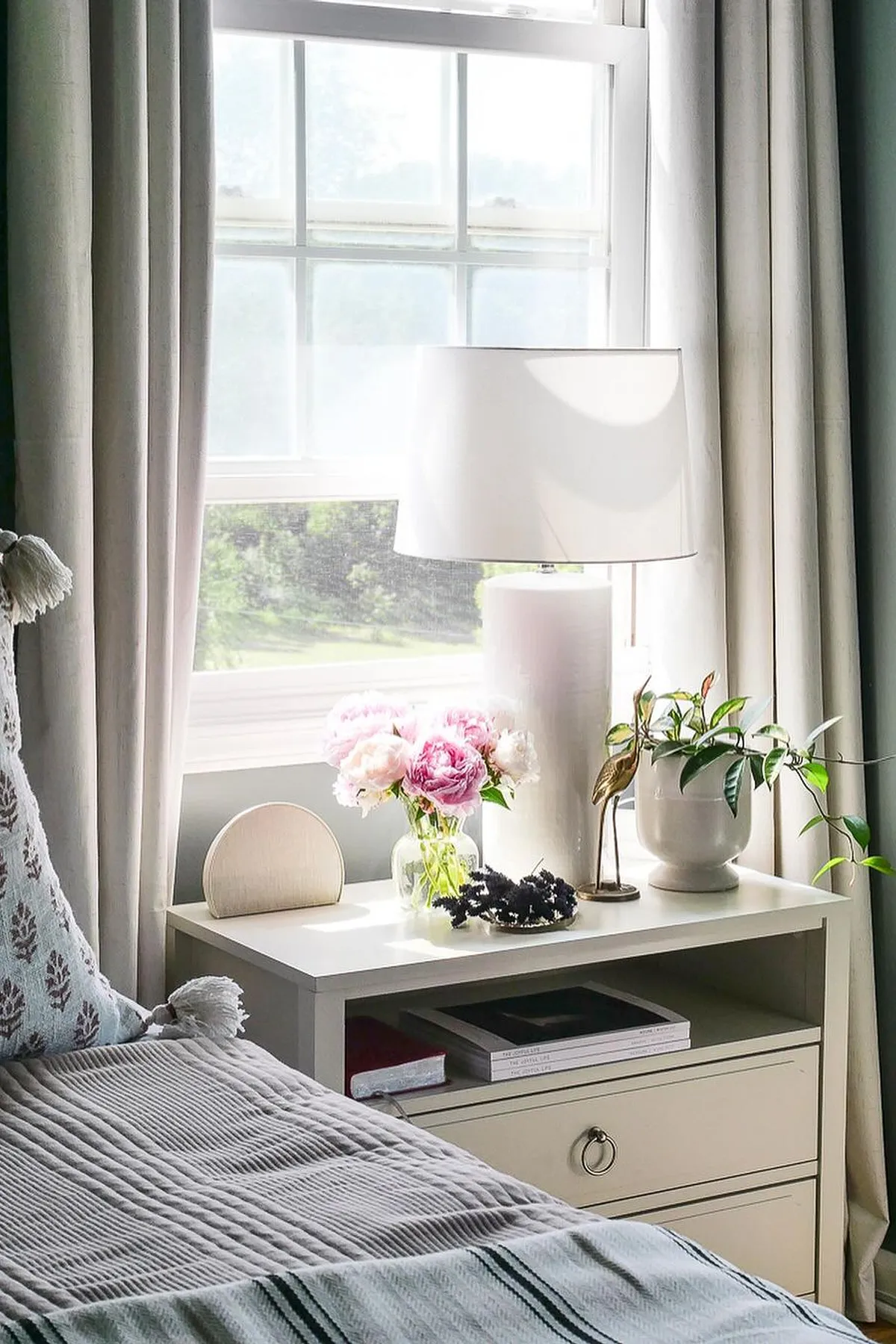 A serene bedroom with a white bed, patterned bedding, pink flowers on a nightstand, and natural light from a window.