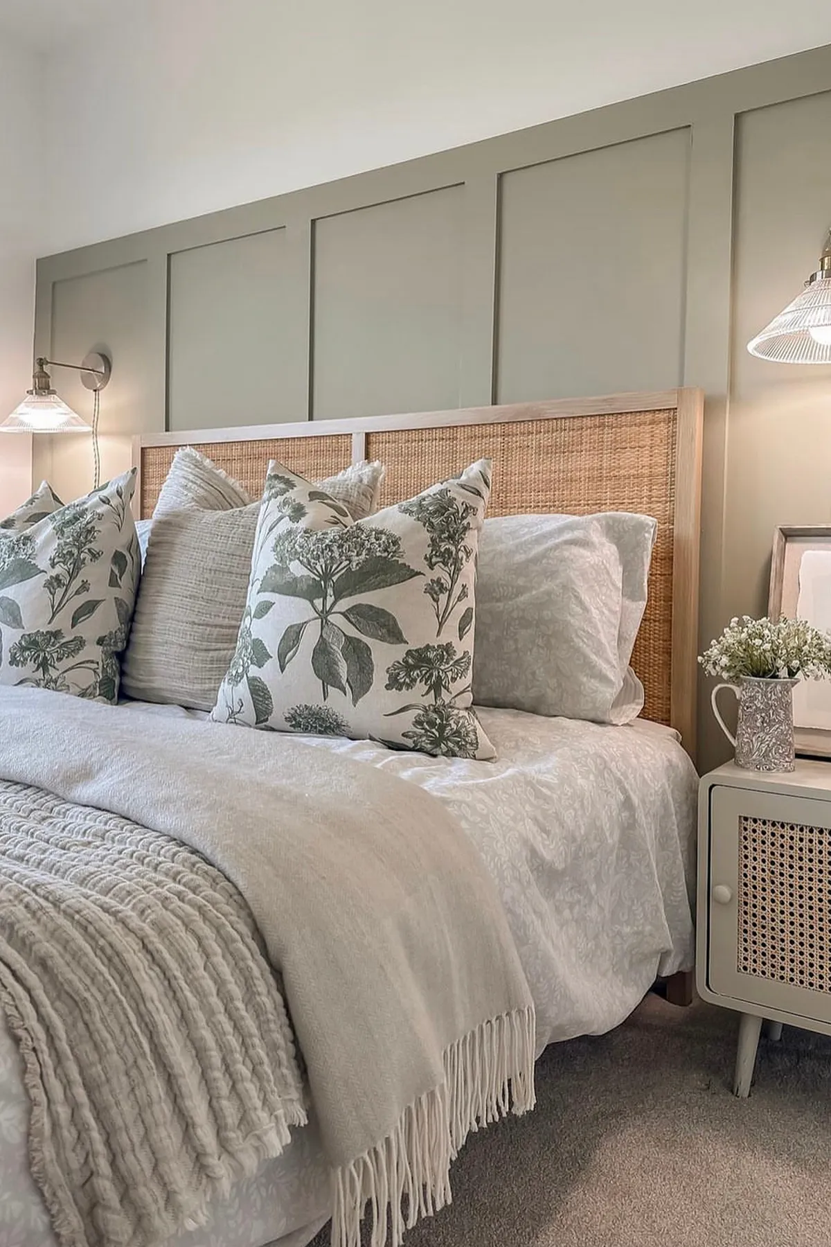 A serene bedroom featuring light bedding, muted green paneling, wall-mounted lamps, and a nightstand with white flowers.