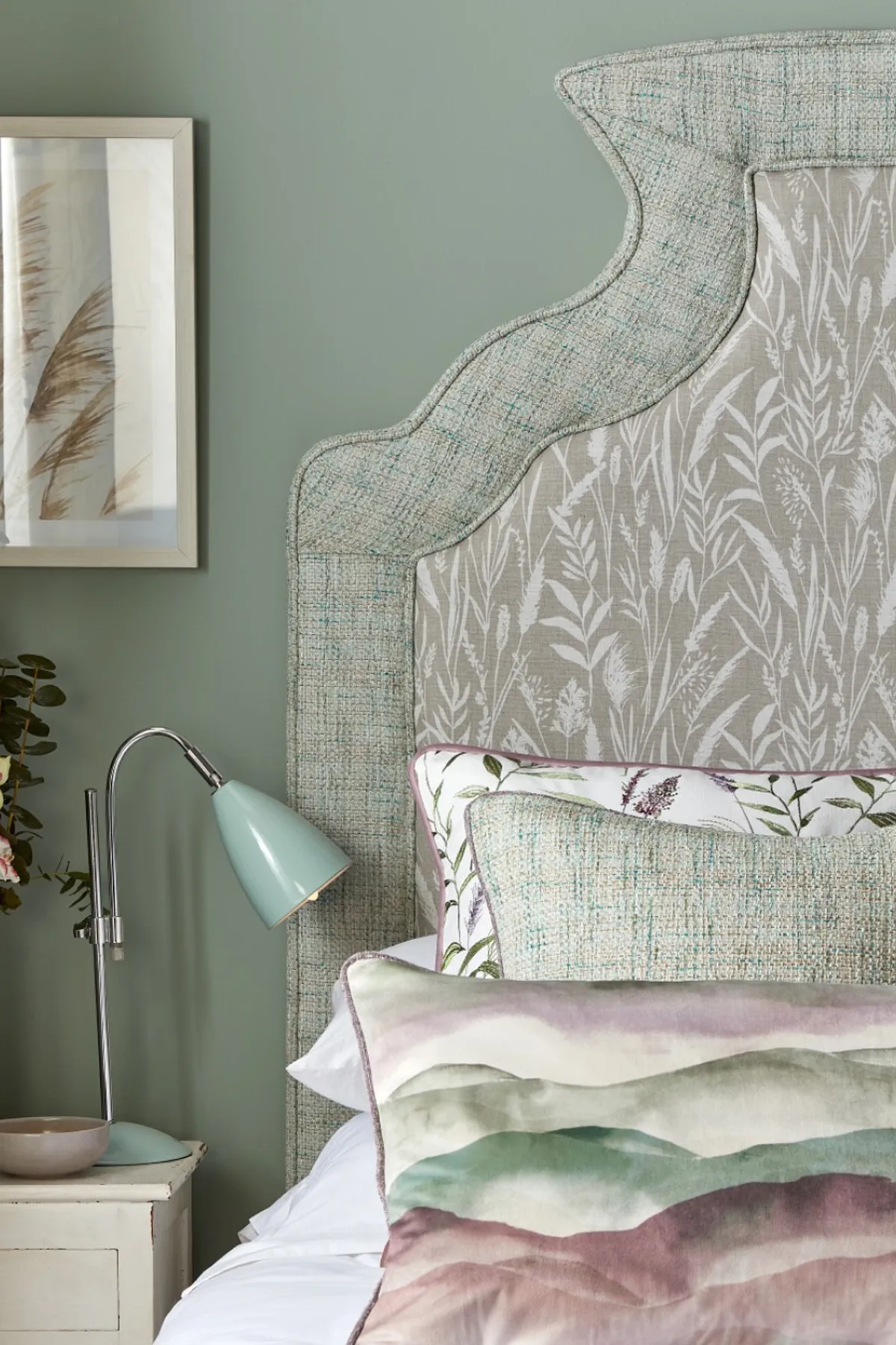 A serene bedroom featuring a floral headboard, decorative pillows, framed plant art, fresh flowers, and a teal lamp.