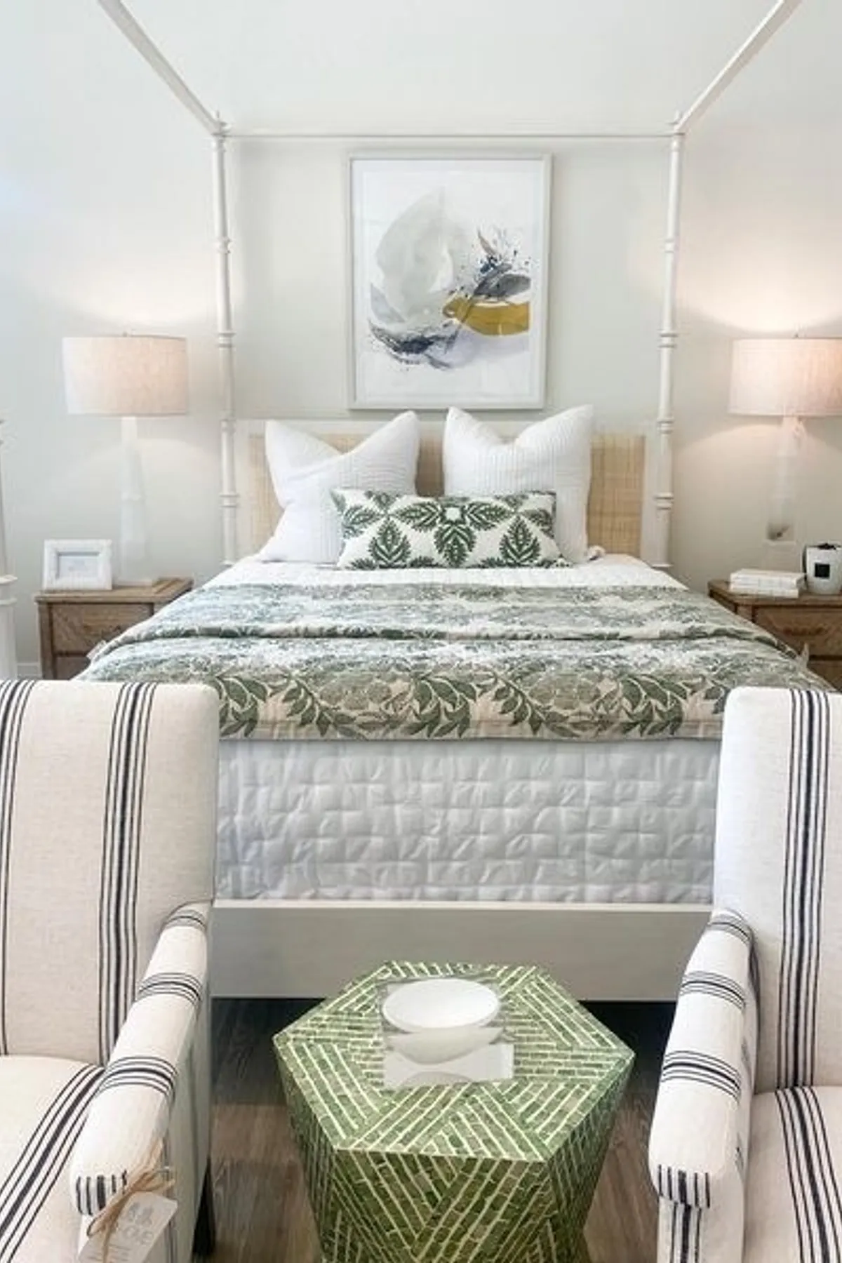 A bright bedroom featuring a four-poster bed, white bedding, patterned pillows, abstract art, wooden nightstands, and a green hexagonal stool.