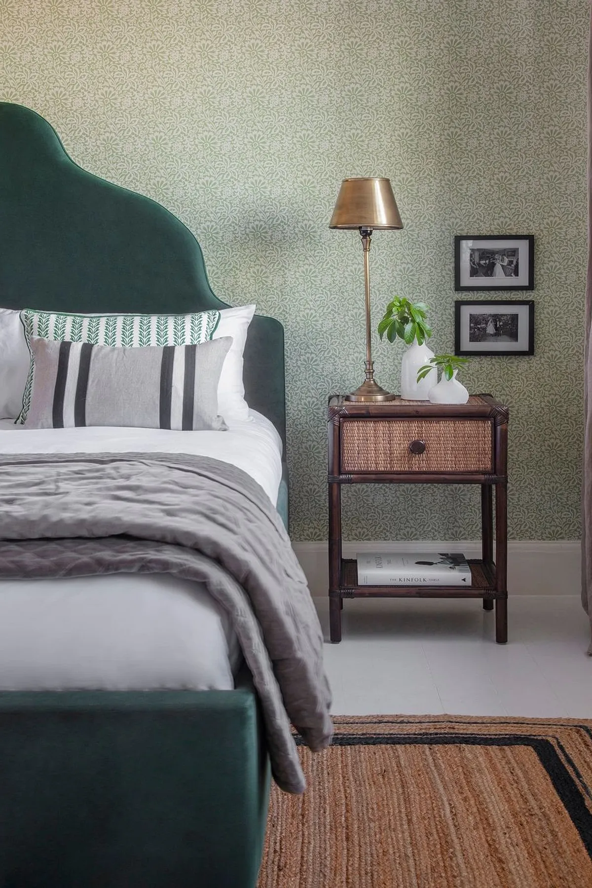 A stylish bedroom featuring a green upholstered bed, striped pillows, wooden nightstand with brass lamp, and layered beige rugs.