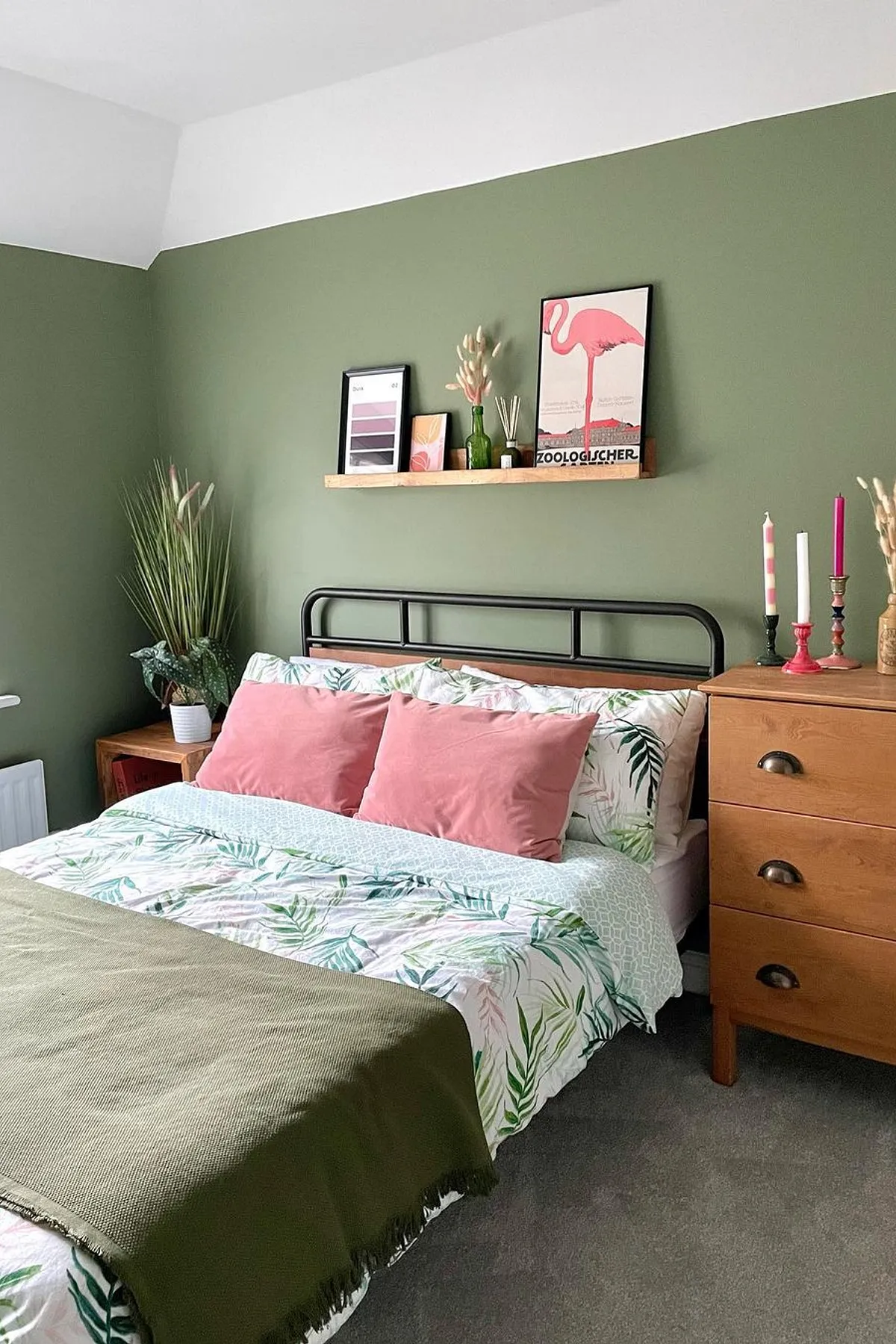 A bedroom featuring a green accent wall, tropical bedding, pink pillows, decorative shelf, and a wooden dresser with candles.
