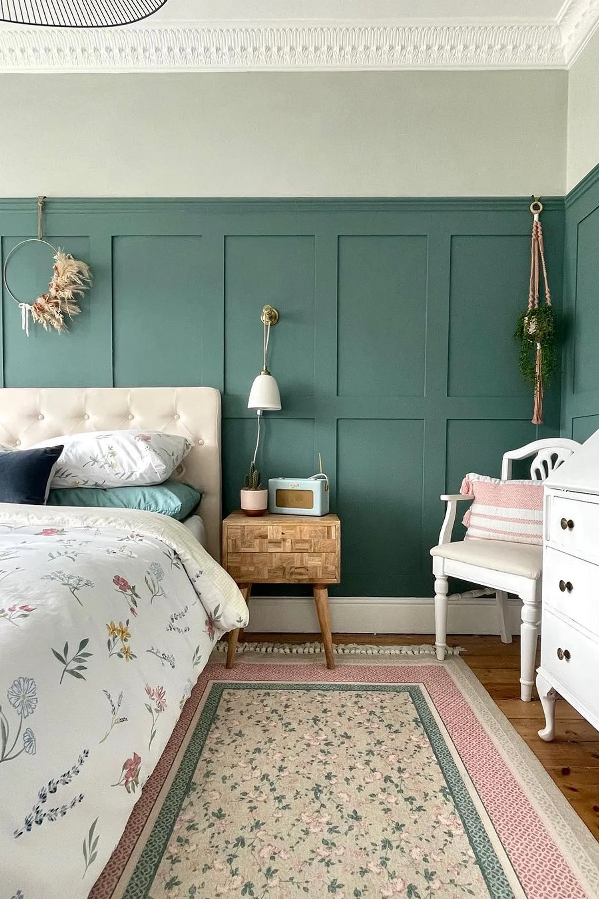 A cozy bedroom with a deep teal wall, floral bedding, vintage dresser, and decorative elements like a wreath and potted plant.