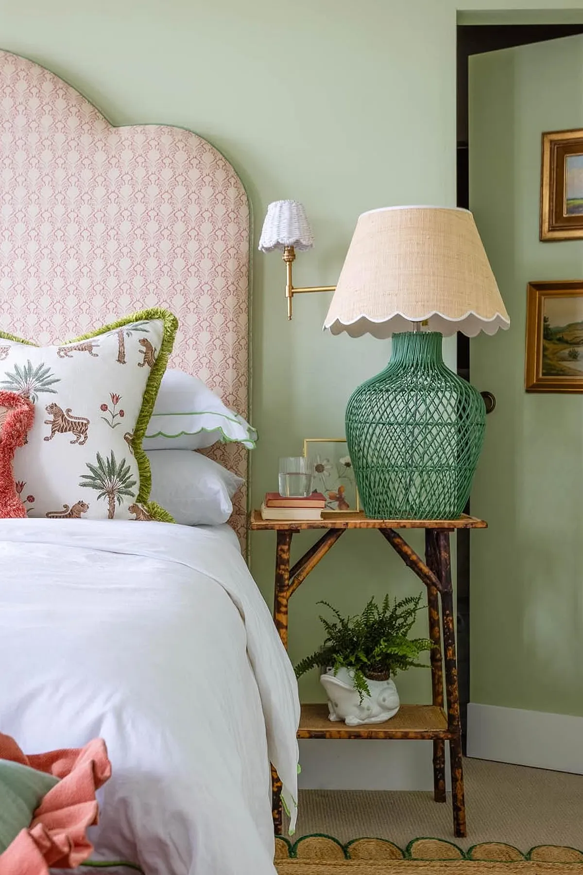 A cozy bedroom with soft green walls, floral pillows, patterned headboard, and decorative elements like a lamp and framed art.