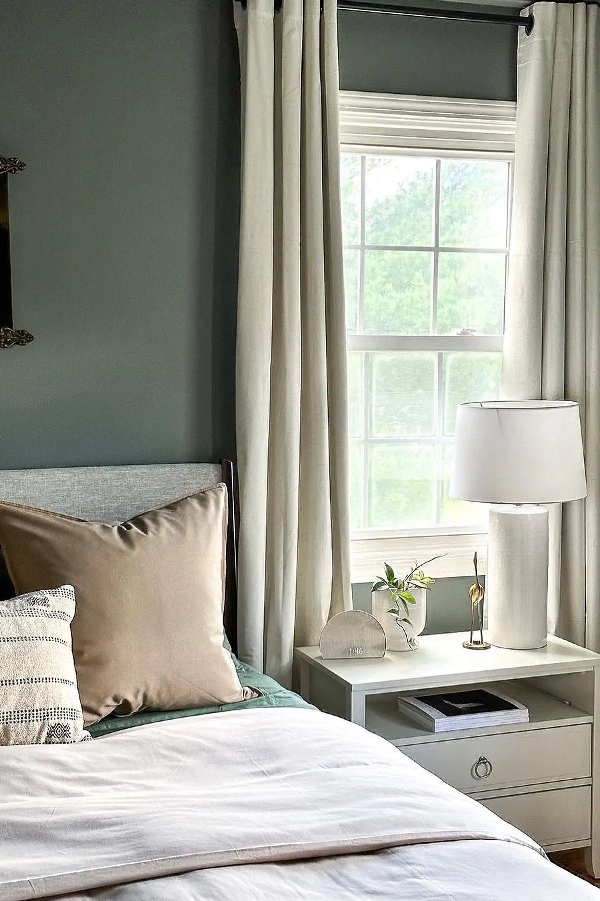 A serene bedroom with muted green walls, beige and white bedding, and a white nightstand topped with decorative items and a plant.