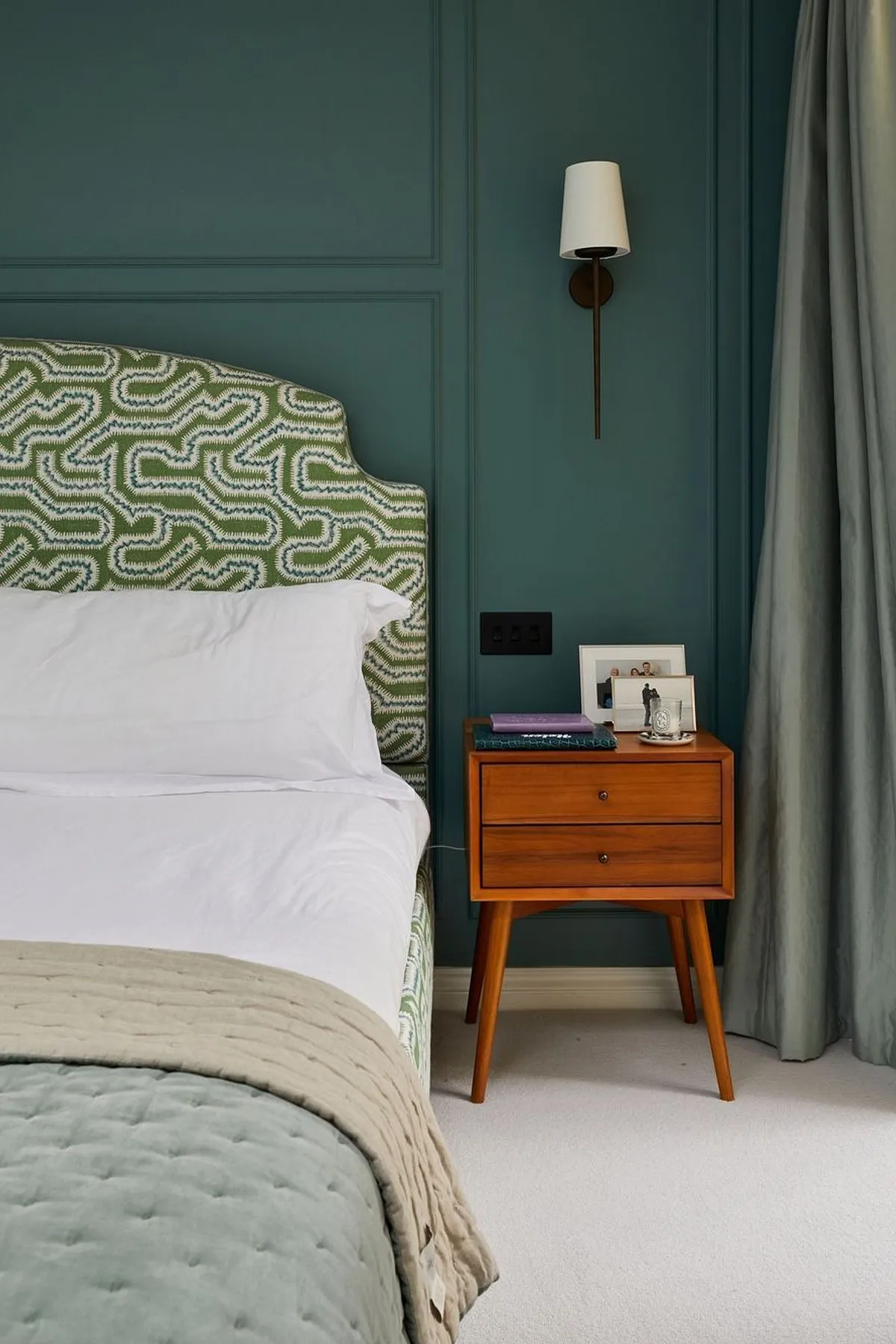A bedroom with deep teal walls, geometric headboard, wooden nightstand, and a wall-mounted lamp beside a window view of trees.