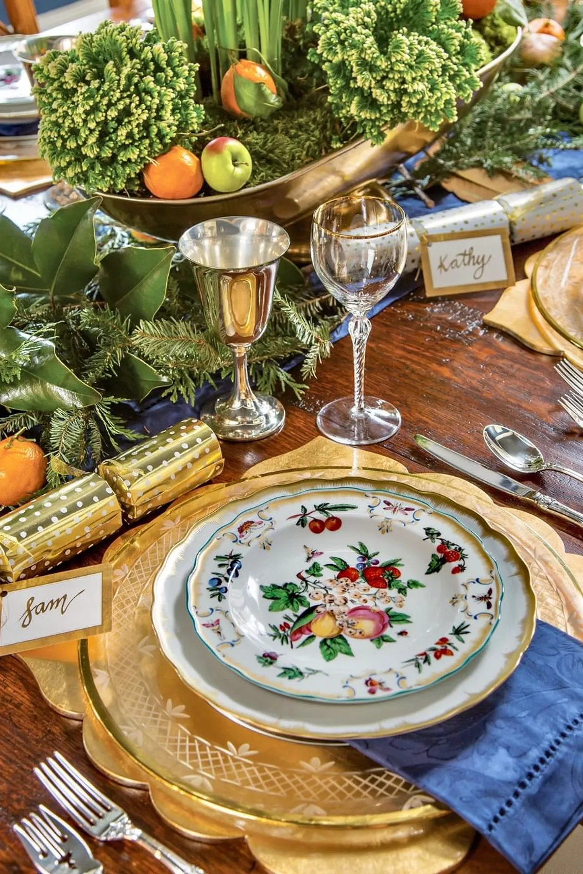 Dining table featuring vintage and modern tableware, golden charger, decorative fruit bowl, and personalized name tags for a festive look.