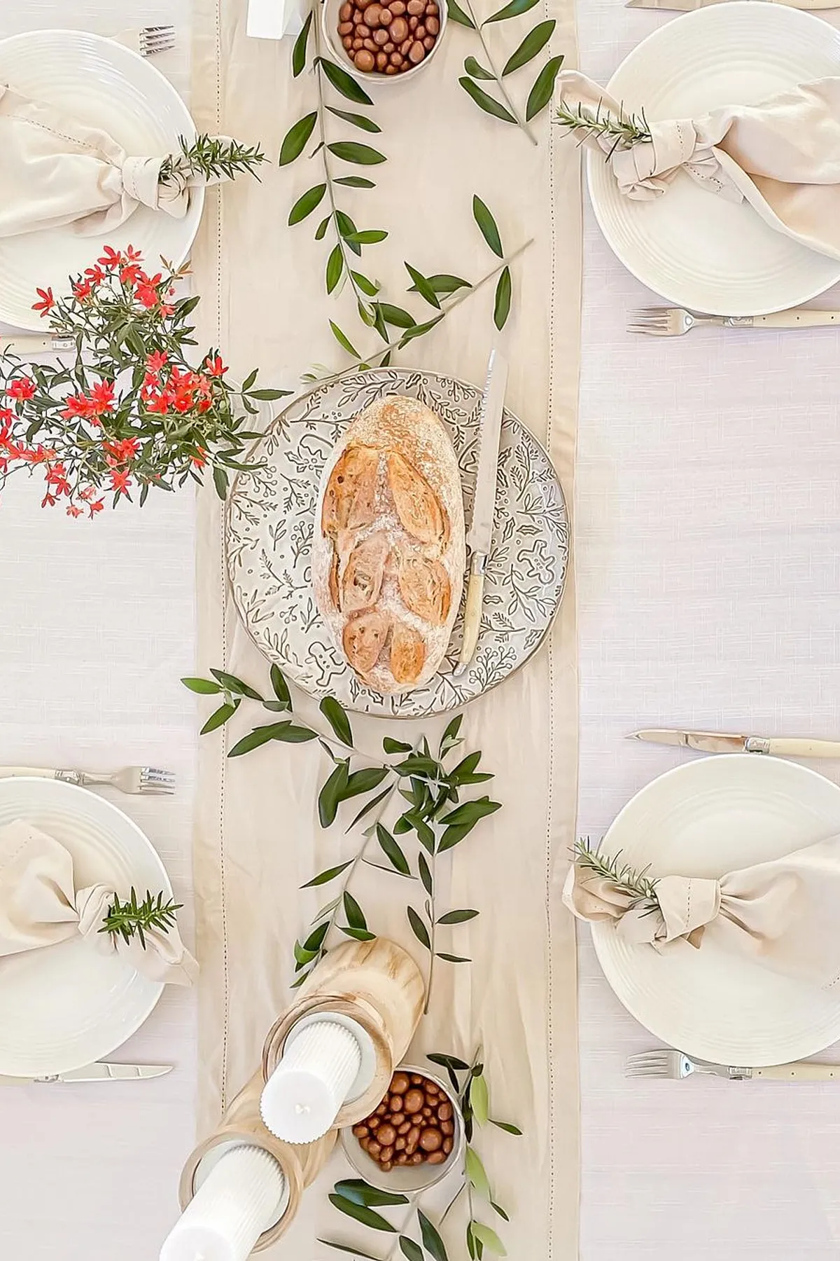 Dining table set with rustic bread, candles, white plates, almonds, greenery, and red flowers for a warm and inviting look.