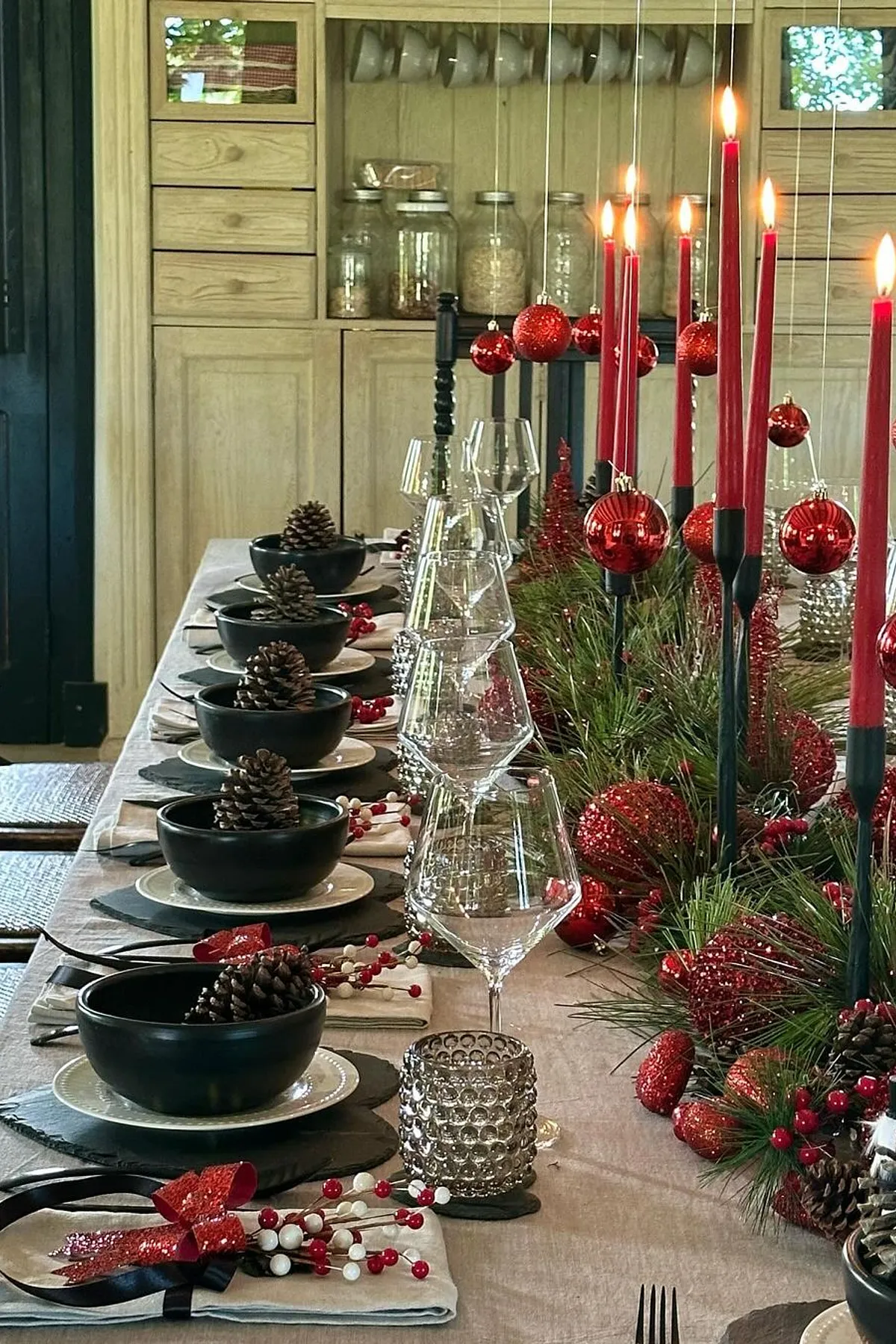 Dining table decorated with hanging red candles, black bowls with pine cones, and red ornaments for a warm festive ambiance.