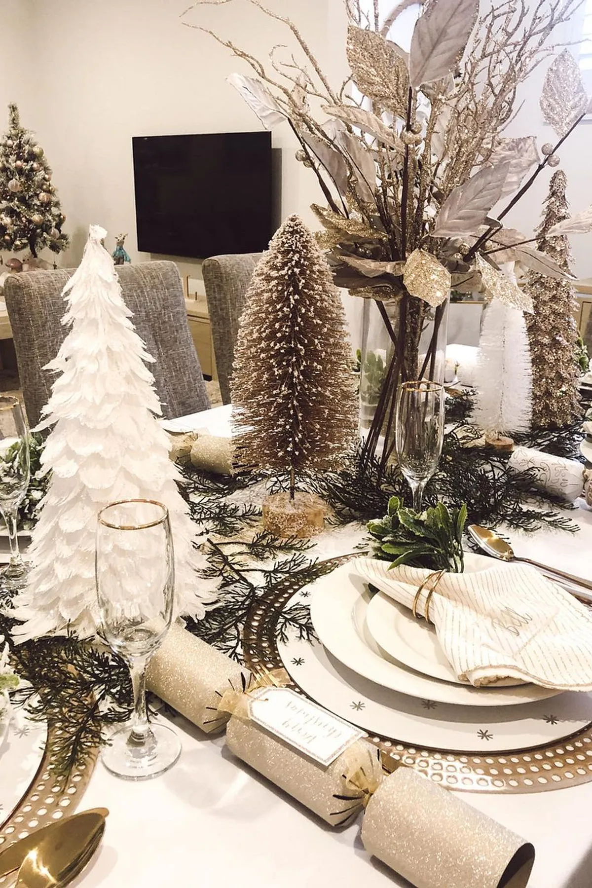 Dining table with white and gold decor, featuring a feathered tree centerpiece, personalized crackers, dried leaves, and a Christmas tree backdrop.
