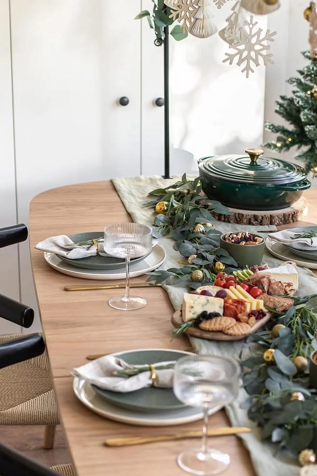 Dining table with green runner featuring fruits, cheeses, and crackers, surrounded by elegant chairs and decorated with snowflakes and stars.
