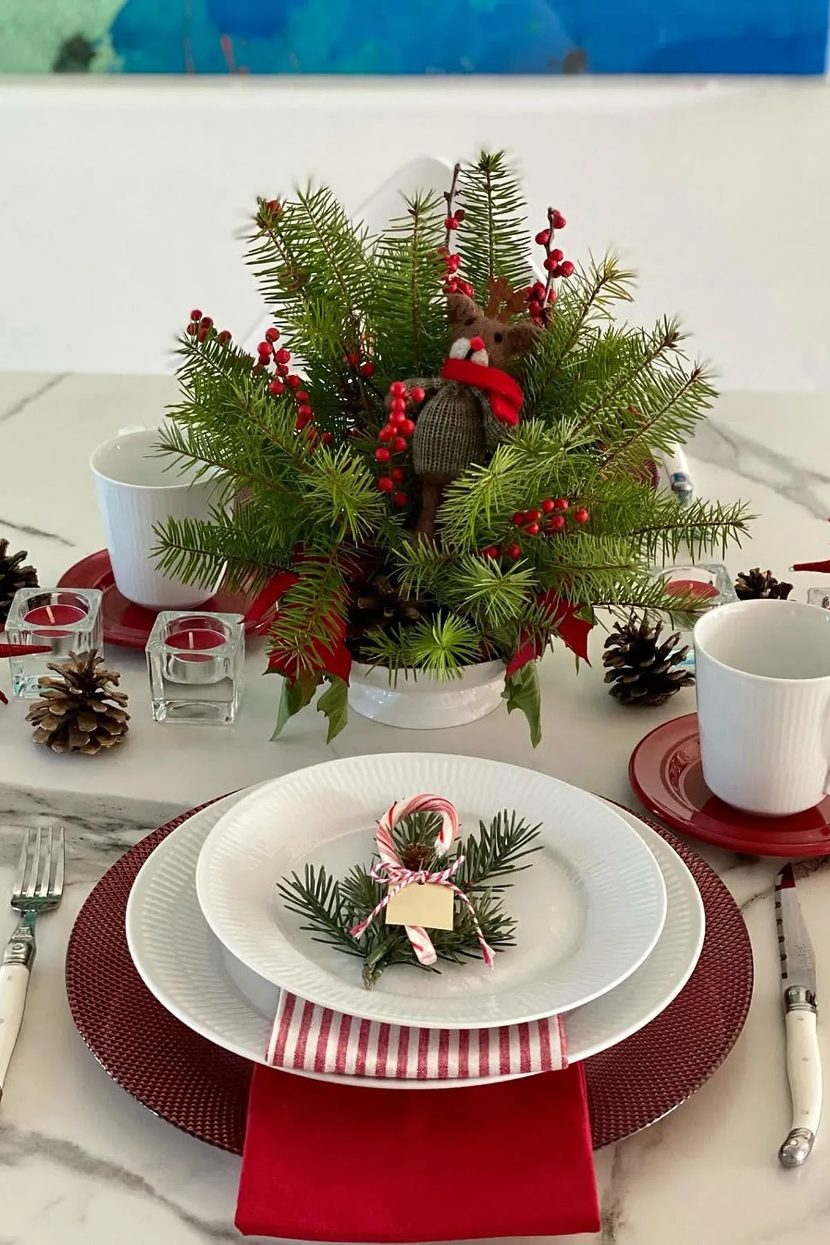 Festive holiday table with evergreen centerpiece, red berries, plush reindeer, white cups, red saucer, and pinecones on a marble surface.