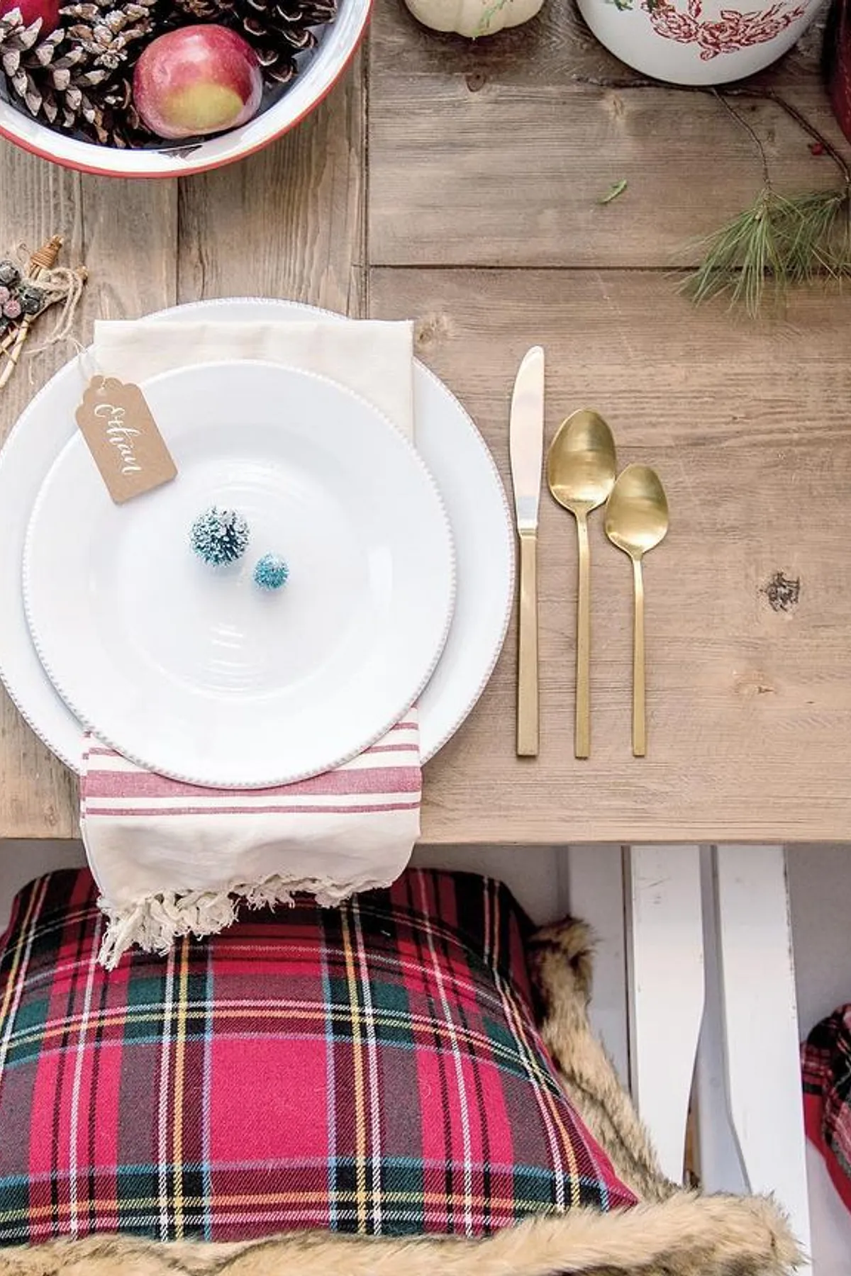Rustic wooden table set with white plates, golden utensils, apples, pinecones, and plaid cushions in warm earthy tones.