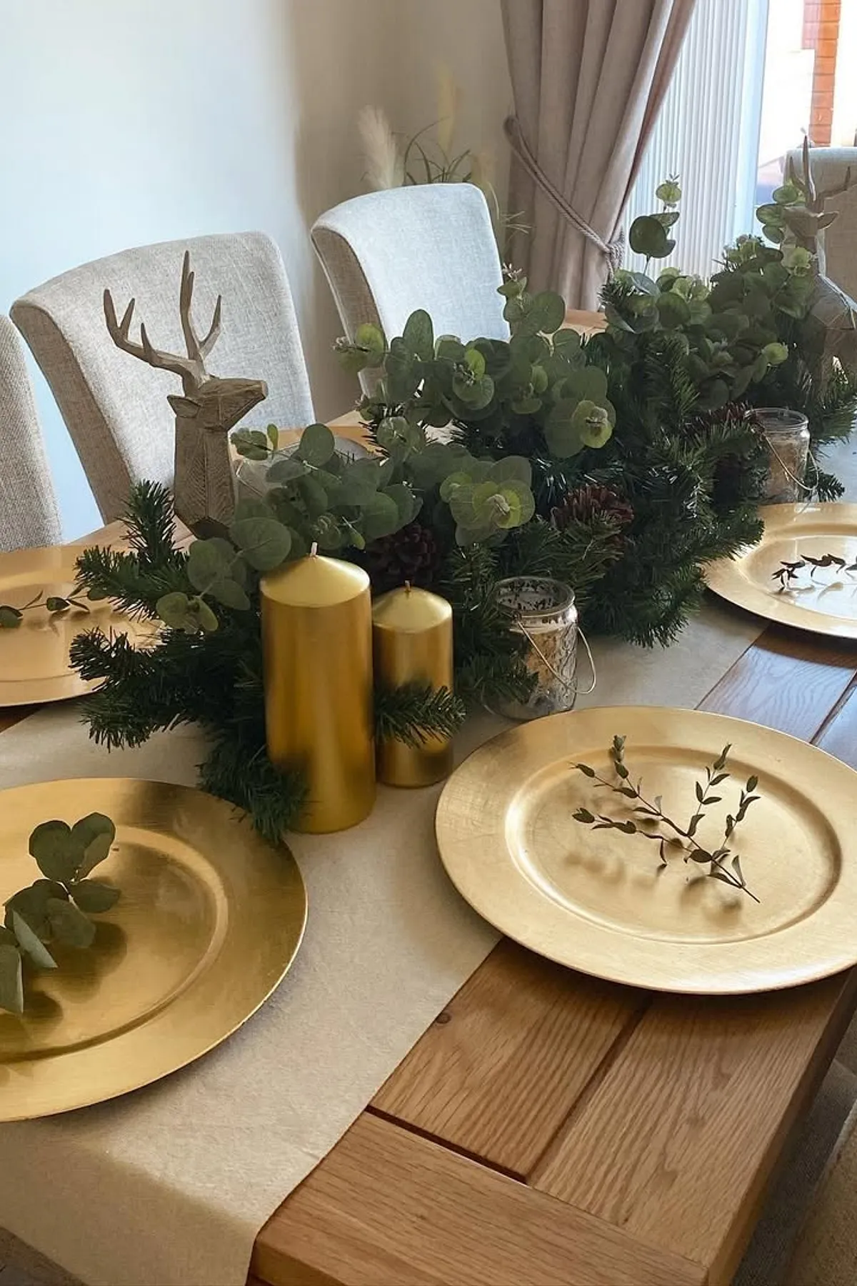Dining table with golden charger plates, greenery centerpiece, tall candles, beige chairs, and a view of a brick wall.
