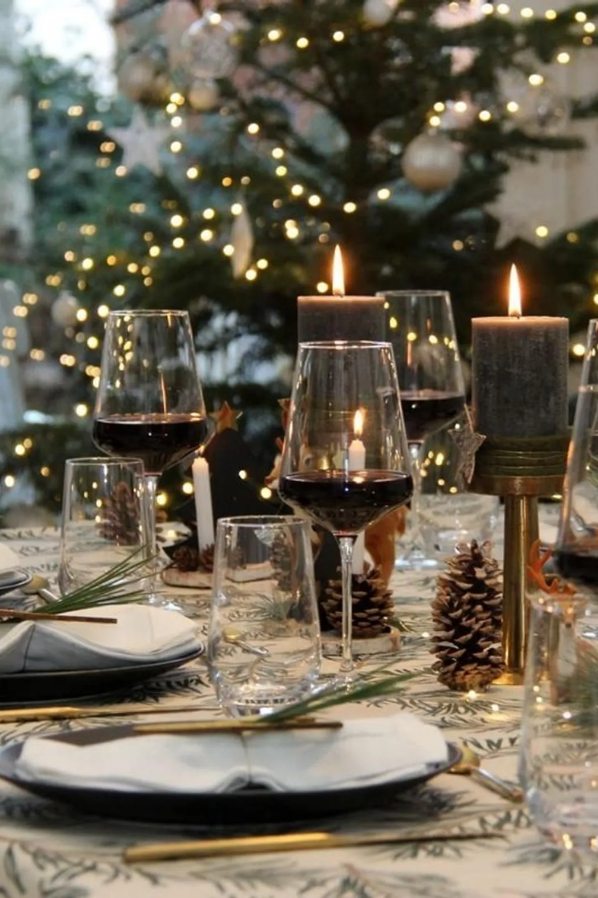 Dining table with patterned tablecloth, red wine glasses, tall candles, pinecones, and a Christmas tree with fairy lights in the background.