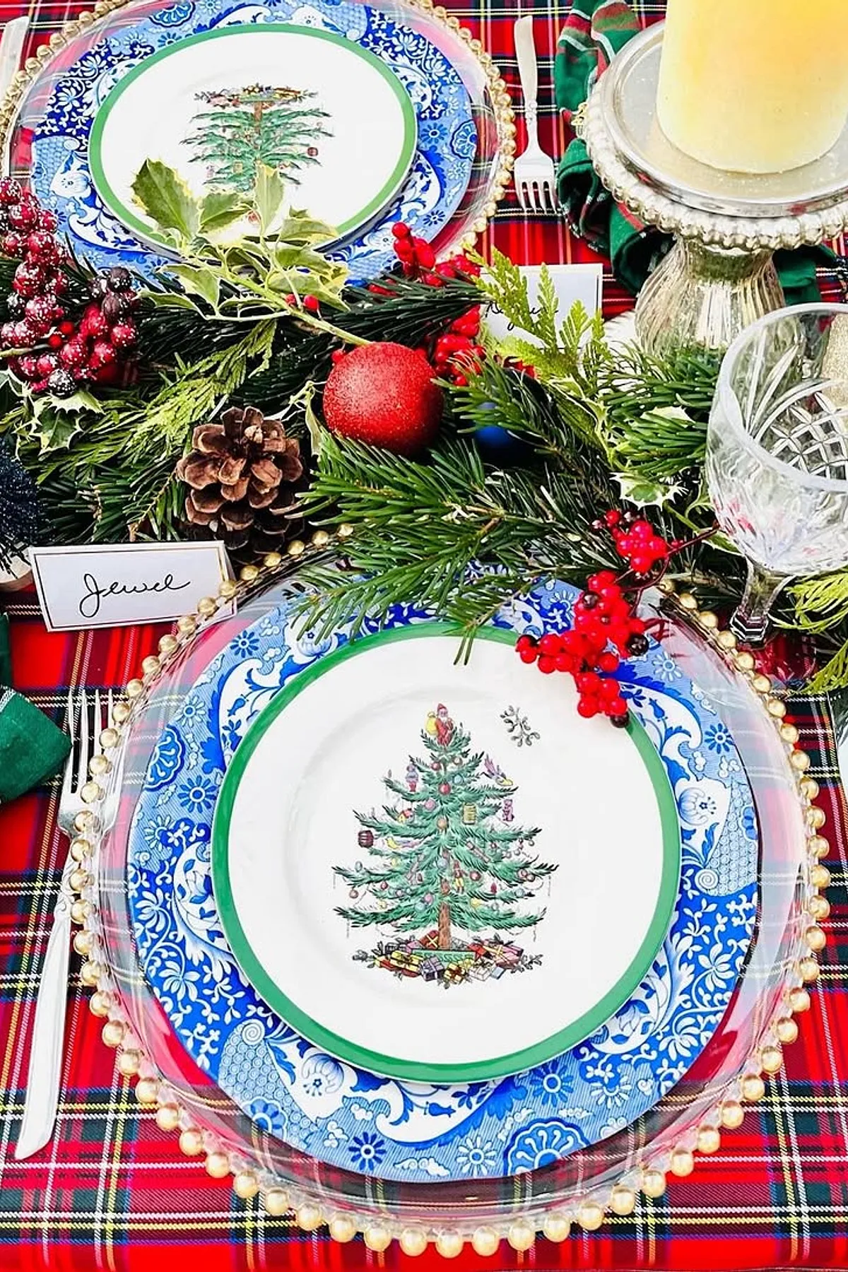 Festive table with plaid tablecloth, blue and white plates, green napkins, candles, pine cones, and holiday decorations for Christmas meal.