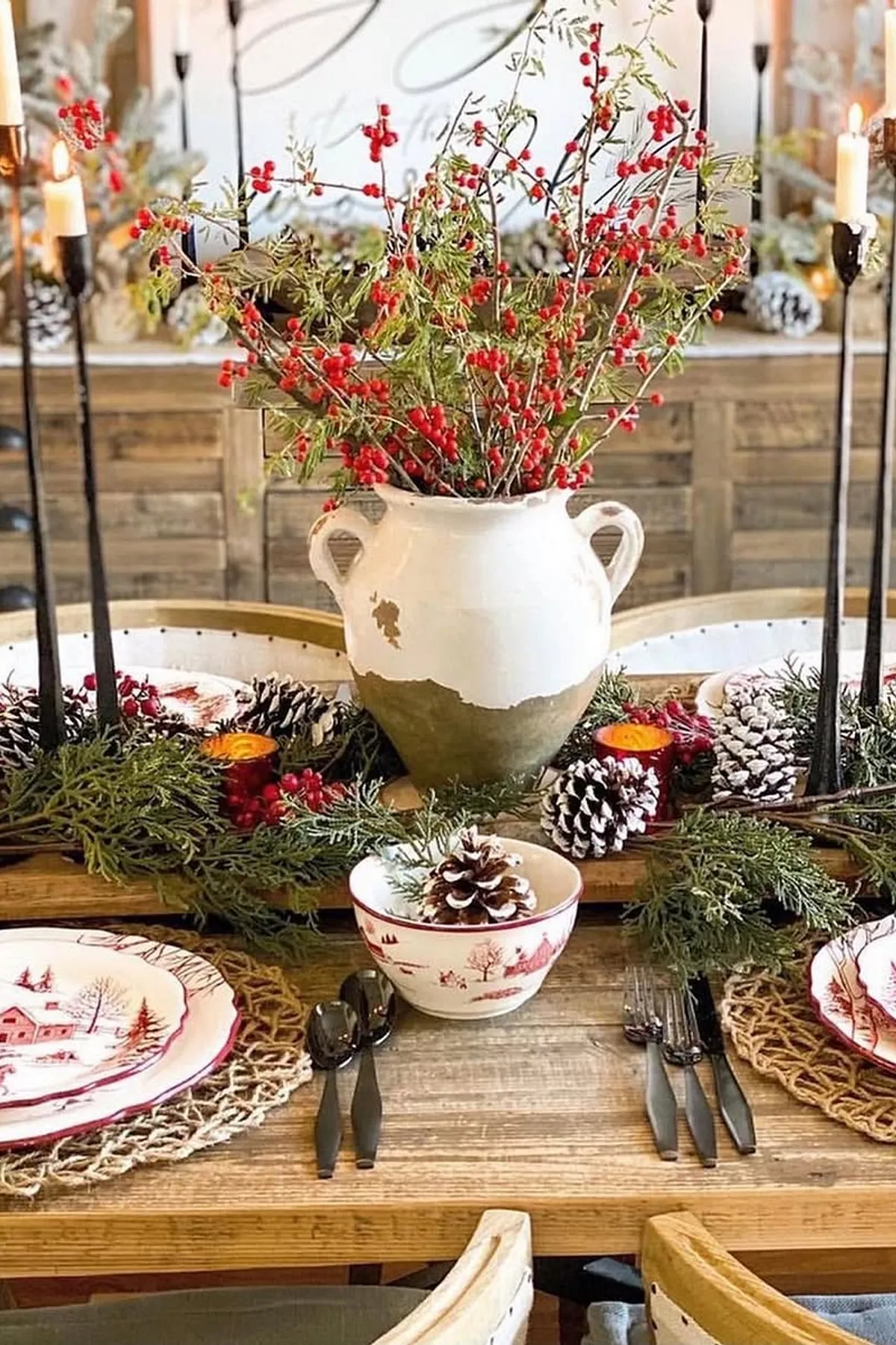 Festive dining table with rustic vase, red berries, candles, winter scene plates, pinecones, woven placemats, and holiday decorations in background.