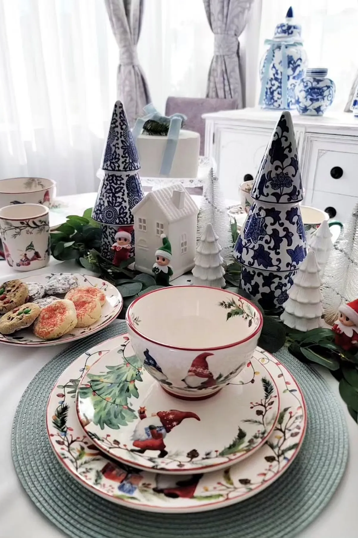 Festive table setting with ceramic plates, gnome designs, porcelain trees, elf figurines, cookies, and pastries under natural light.