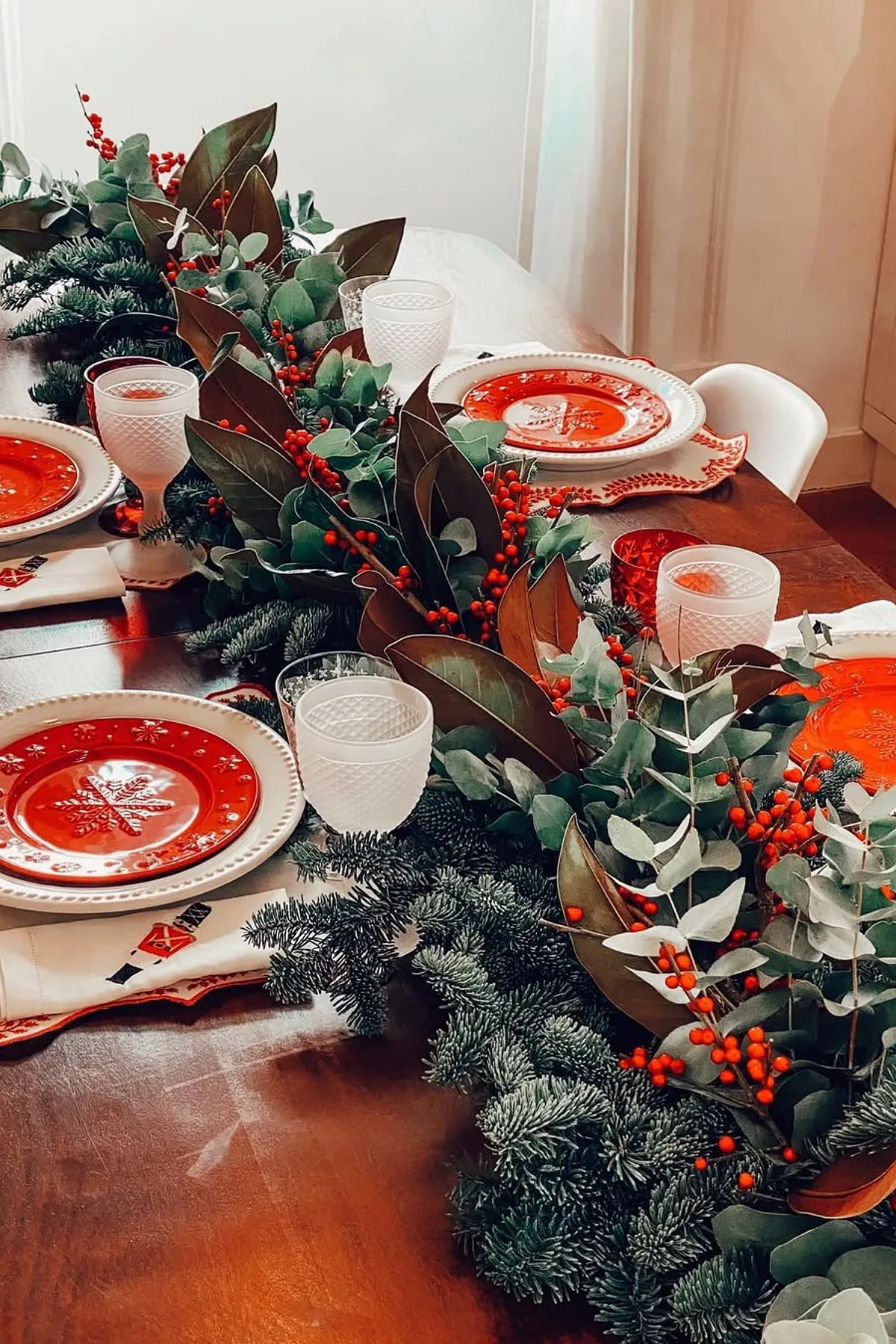 Festive dining table with eucalyptus garland, red plates, white wine glasses, and soft light filtering through white curtains.