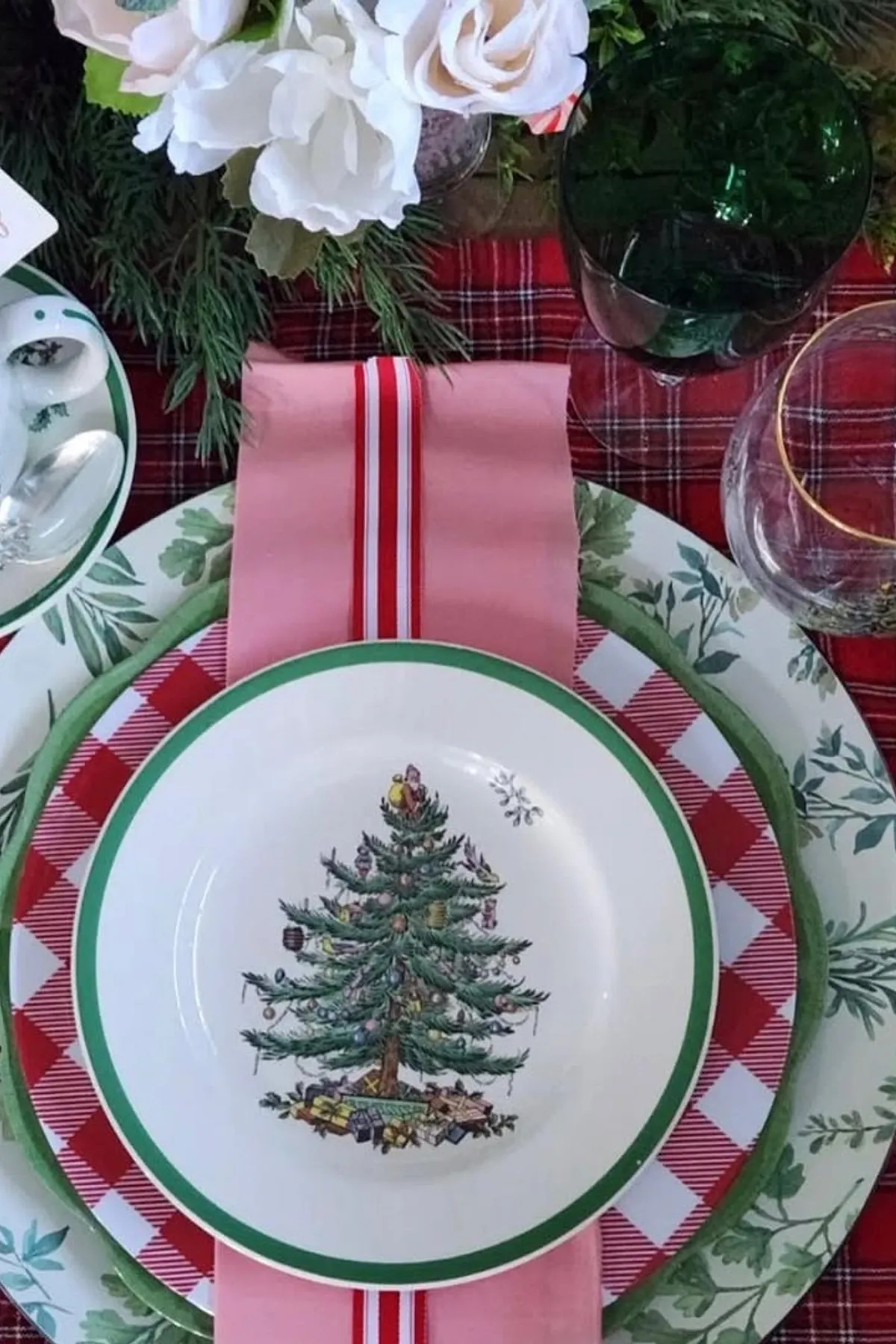 Festive table setting with red and green decor, Christmas tree plate, candy cane cup, silverware, and white roses in a vase.